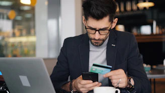 a man sitting at a table looking at his cell phone and holding a credit card