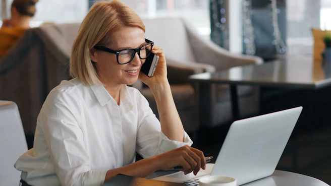 an older woman with black frame glasses talking on a cell phone while using a laptop
