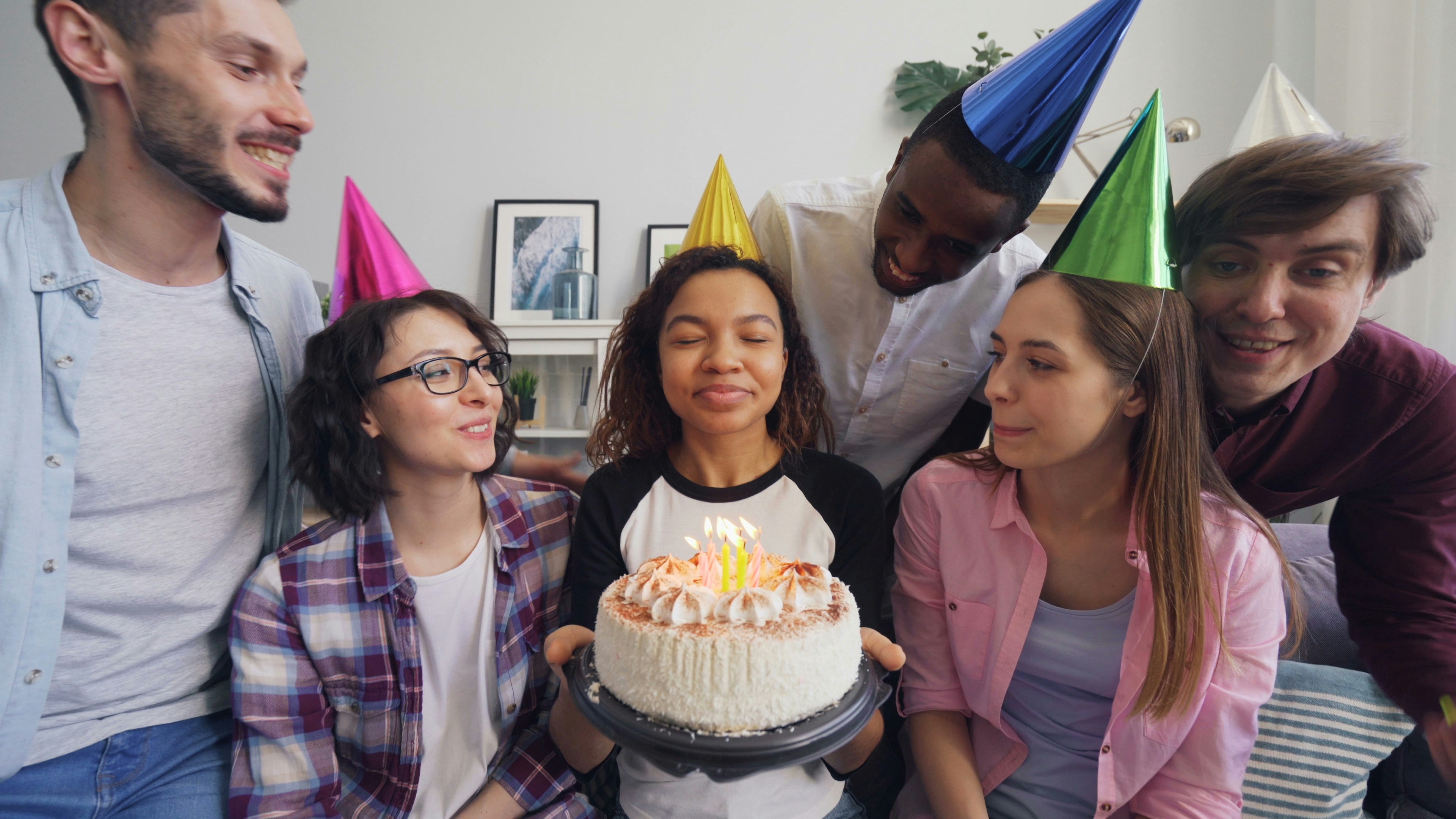 Friends in party hats round a woman holding a birthday cake