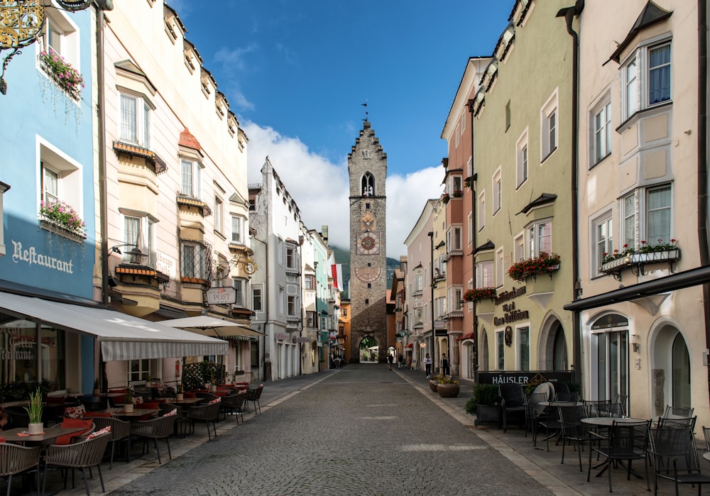 a narrow street with tables and chairs and a clock tower in the background