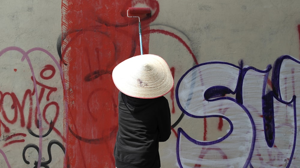 a person with a hat on standing in front of a wall with graffiti
