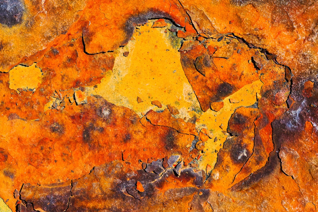 a close up of a rock with yellow paint, Closeup of Rusted Metal Surface With Orange and Brown Patterns - A close-up image of rusted metal, showing a complex pattern of orange and brown hues. The surface has a rough texture with deep cracks and irregular shapes, indicating years of exposure to the elements. The colors are vibrant and varied, creating a visually interesting abstract design.