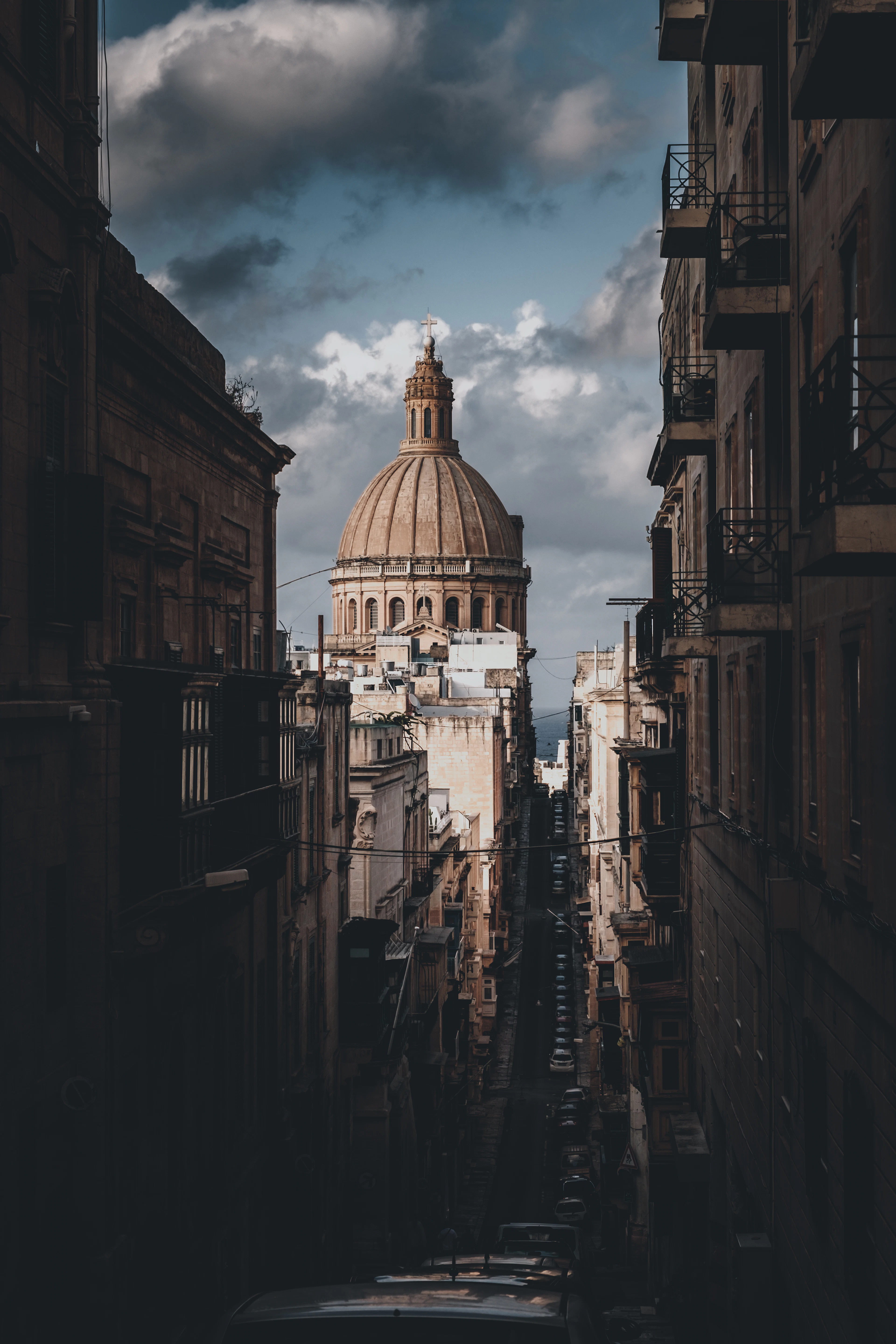 The great stone dome of a Valletta church rising at the end of a steep, shadowed side street lined with balconied townhouses, under heavy storm clouds.