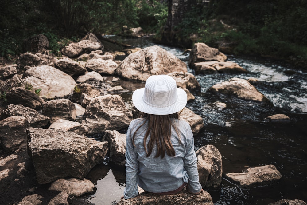 a woman in a white hat is sitting on rocks by a stream