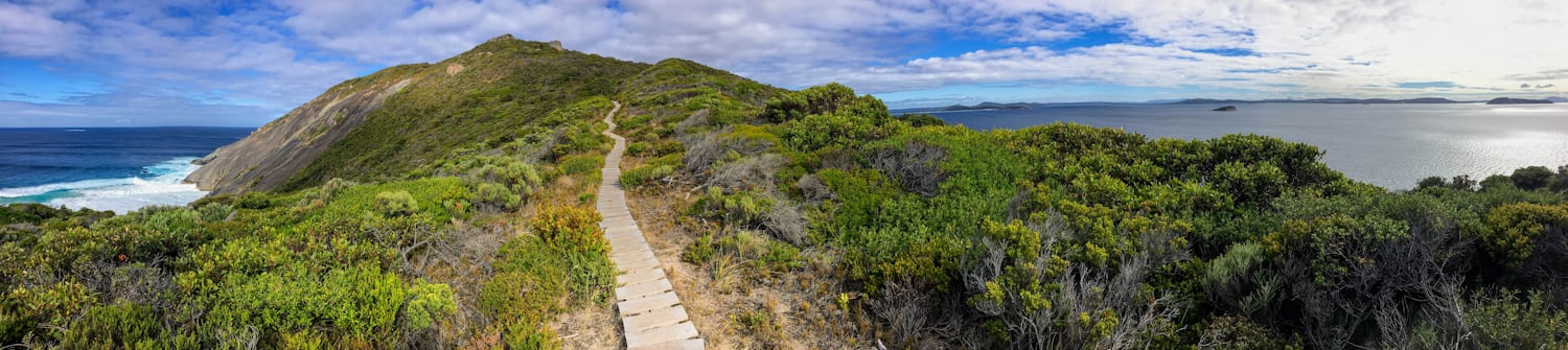 A panorama showing a peninsula covered in green scrub. To the sides, the sea is visible, to the left a rocky escarpment, and to the right various islands. Directly ahead is a wooden path. Above are patches of blue sky showing through cloud.