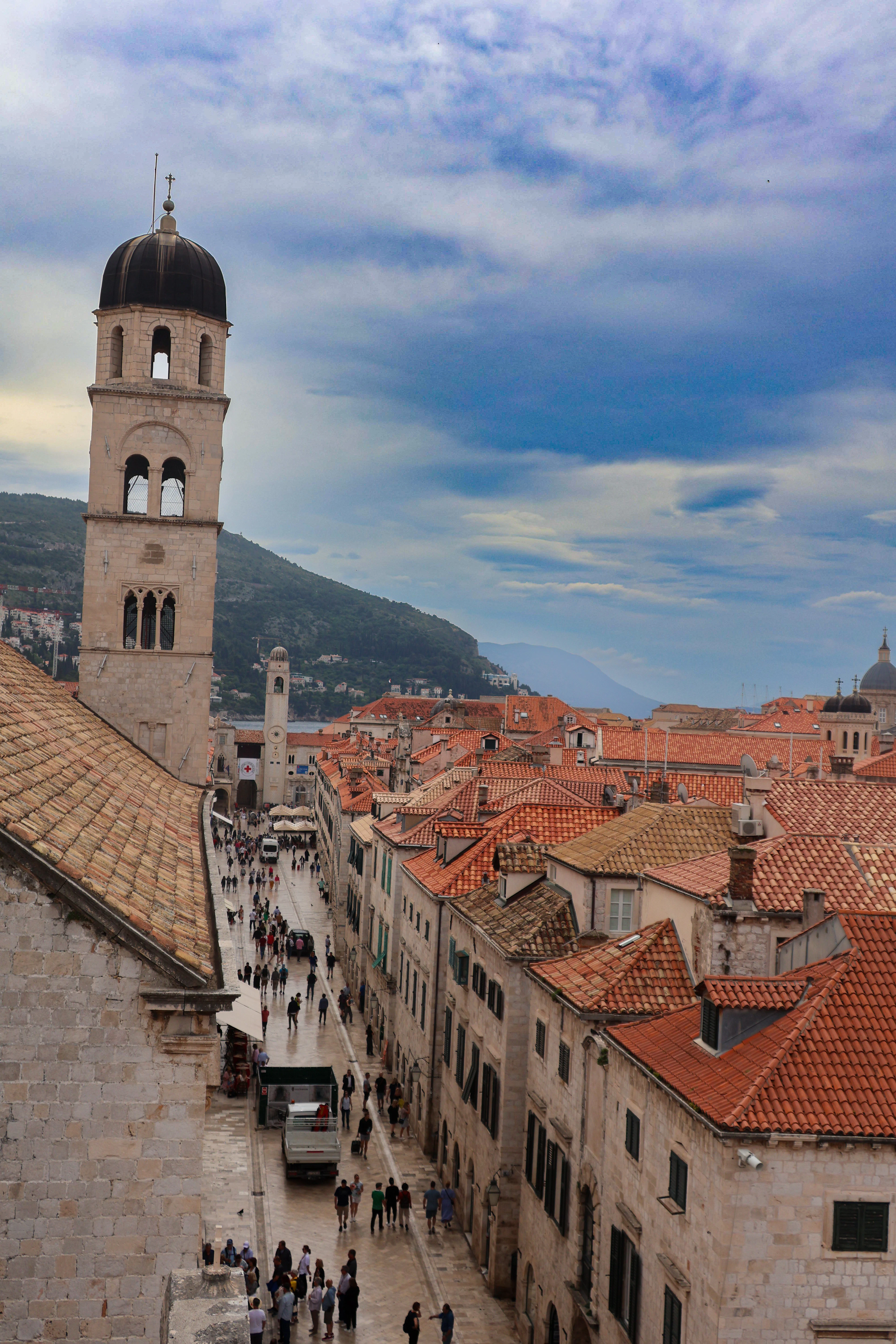 The Stradun seen from high on Dubrovnik's city walls under a bruised blue sky, the Franciscan monastery bell tower rising above the polished limestone street and the tightly packed terracotta roofs of the Old Town.