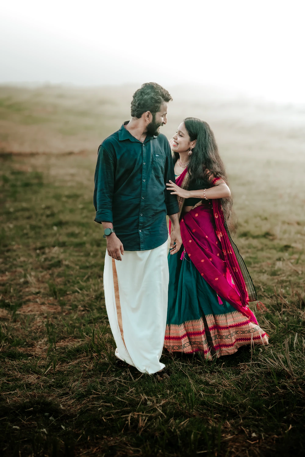 Arjun in a dark green shirt and white mundu with Priya in a magenta and green half saree, in a misty field