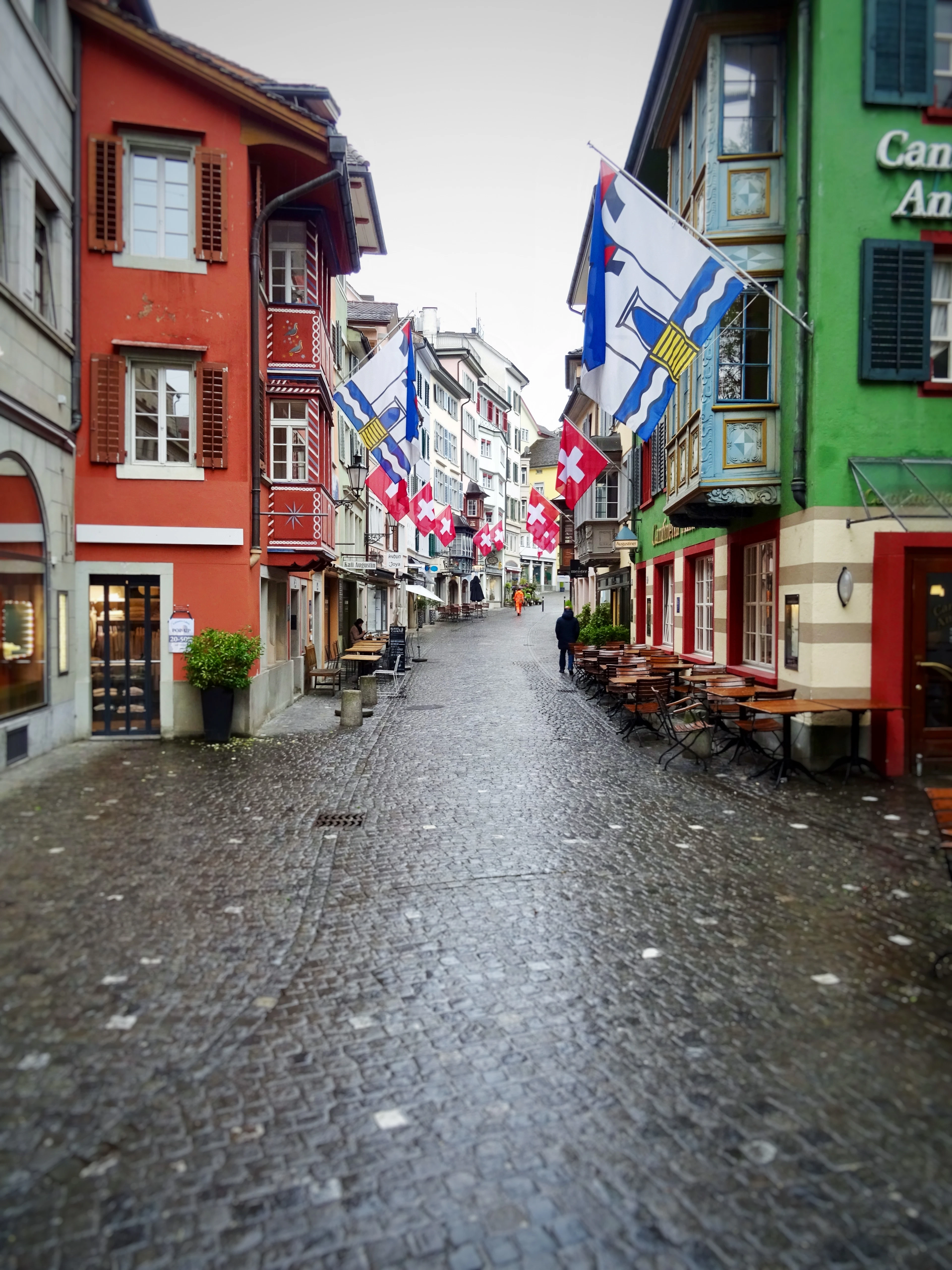 A rain-slicked cobbled lane in Zurich's Old Town hung with Swiss and Zurich cantonal flags, wooden restaurant benches outside red and green painted houses.