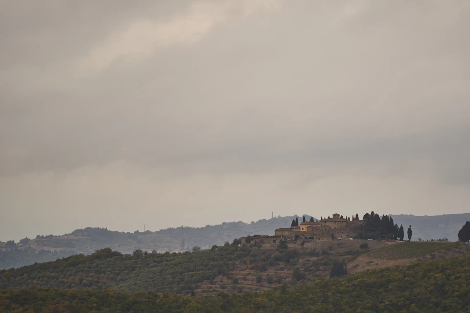 The image depicts a picturesque landscape with a large hill covered in lush green trees and shrubs, nestled between two hills. The sky above is filled with clouds, casting a soft light over the scene. In the distance, there are several buildings visible on the hillside, adding an urban element to the otherwise natural setting.

The image does not contain any discernible text or countable objects, and it focuses solely on the expansive view of the landscape.