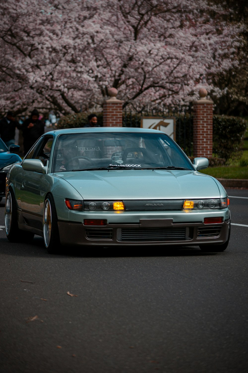 a blue car driving down a street next to a cherry blossom tree