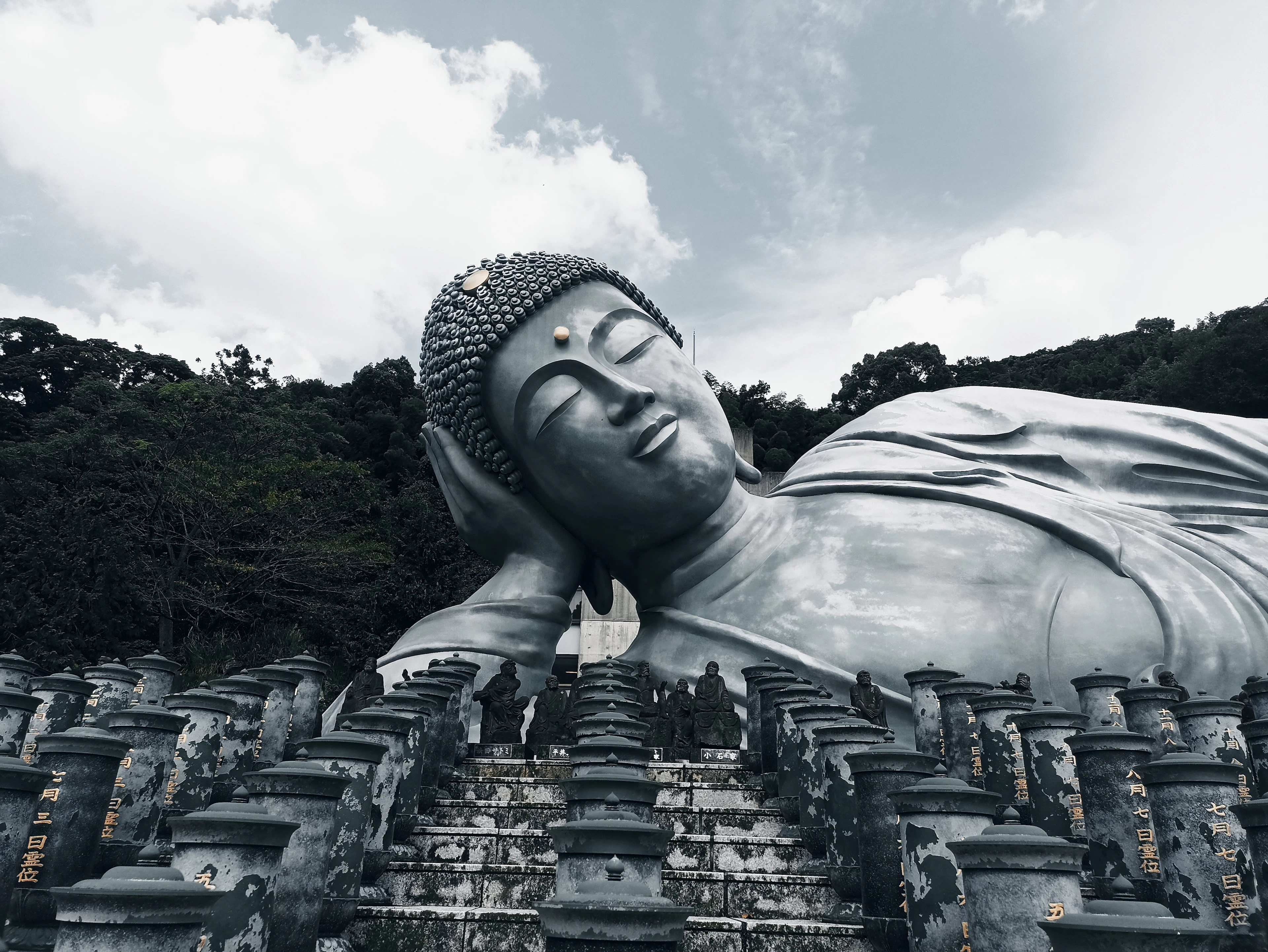 A giant bronze reclining Buddha statue rests on a wooded hillside above rows of stone lanterns.