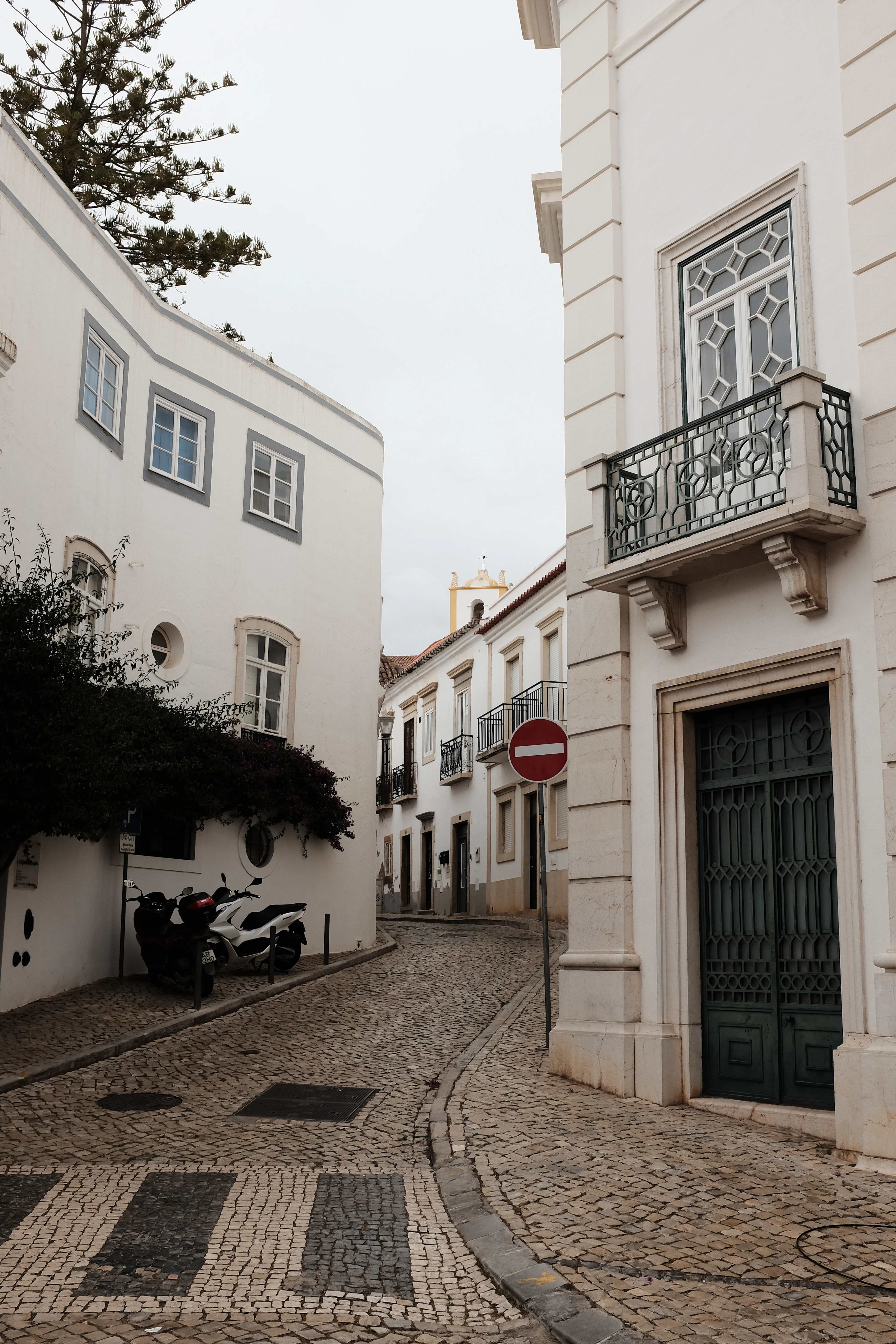 A cobbled street of whitewashed houses in Tavira, Algarve, with wrought-iron balconies, a green studded door and a scooter parked against a wall
