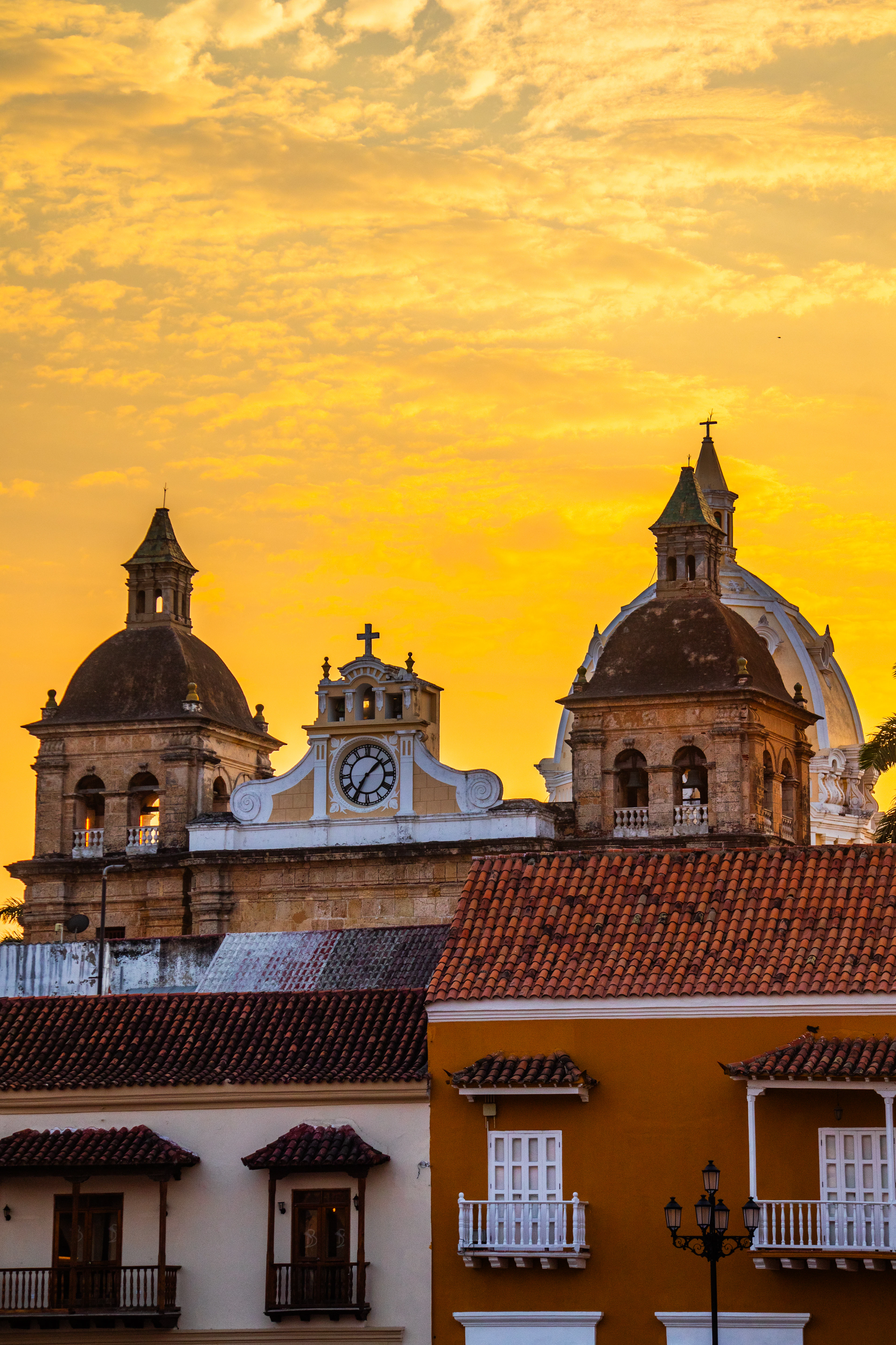 Cartagena's cathedral domes and bell towers are silhouetted against a fiery orange sunset sky.