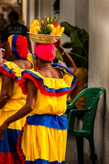 two women in colorful dresses carrying a basket of fruit on their head