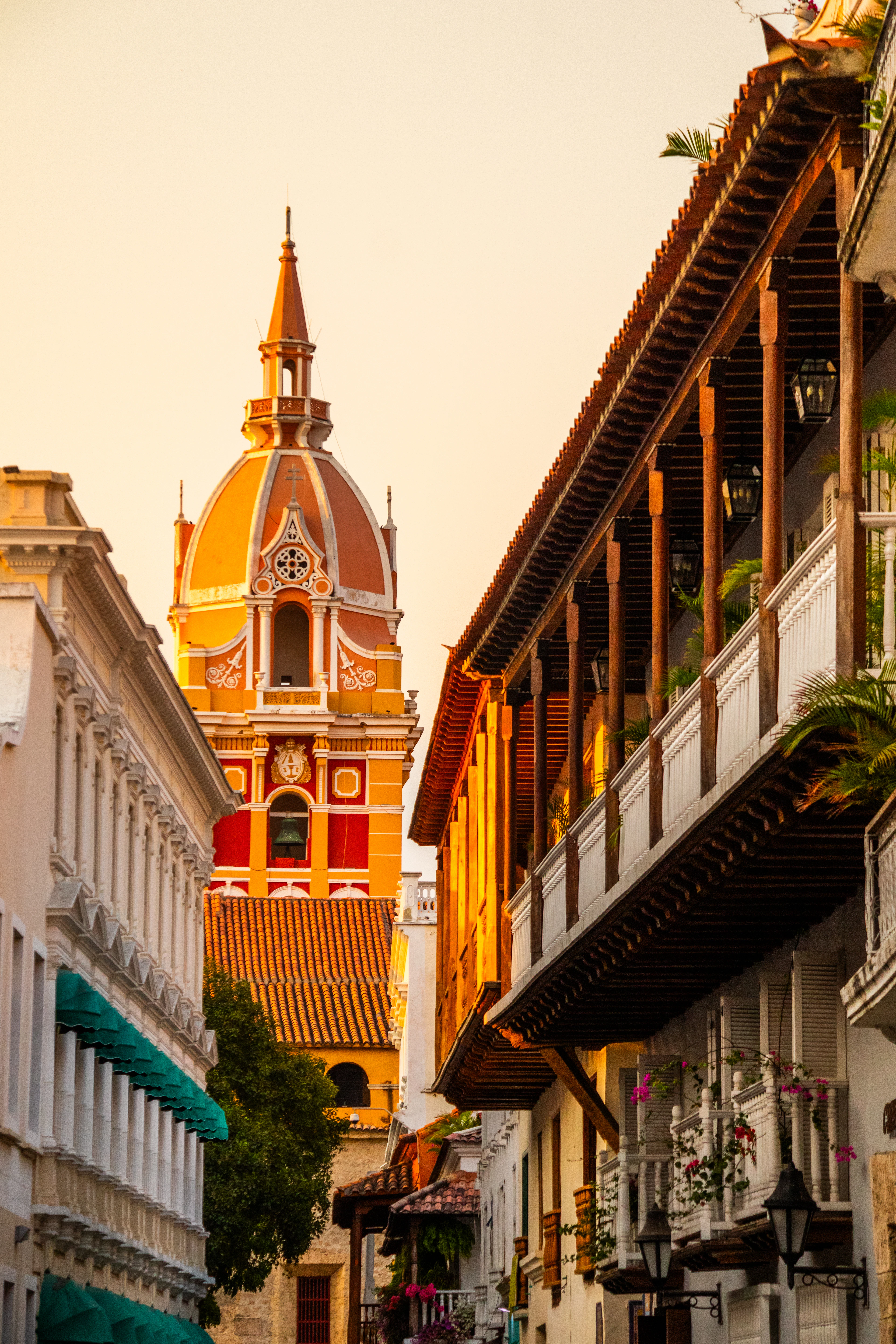 The orange and yellow bell tower and dome of Cartagena's cathedral seen down a lane of wooden colonial balconies at golden hour