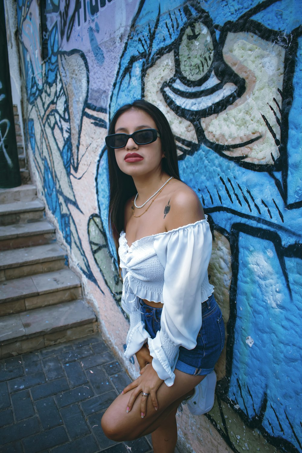 a woman in a white top and sunglasses leaning against a wall