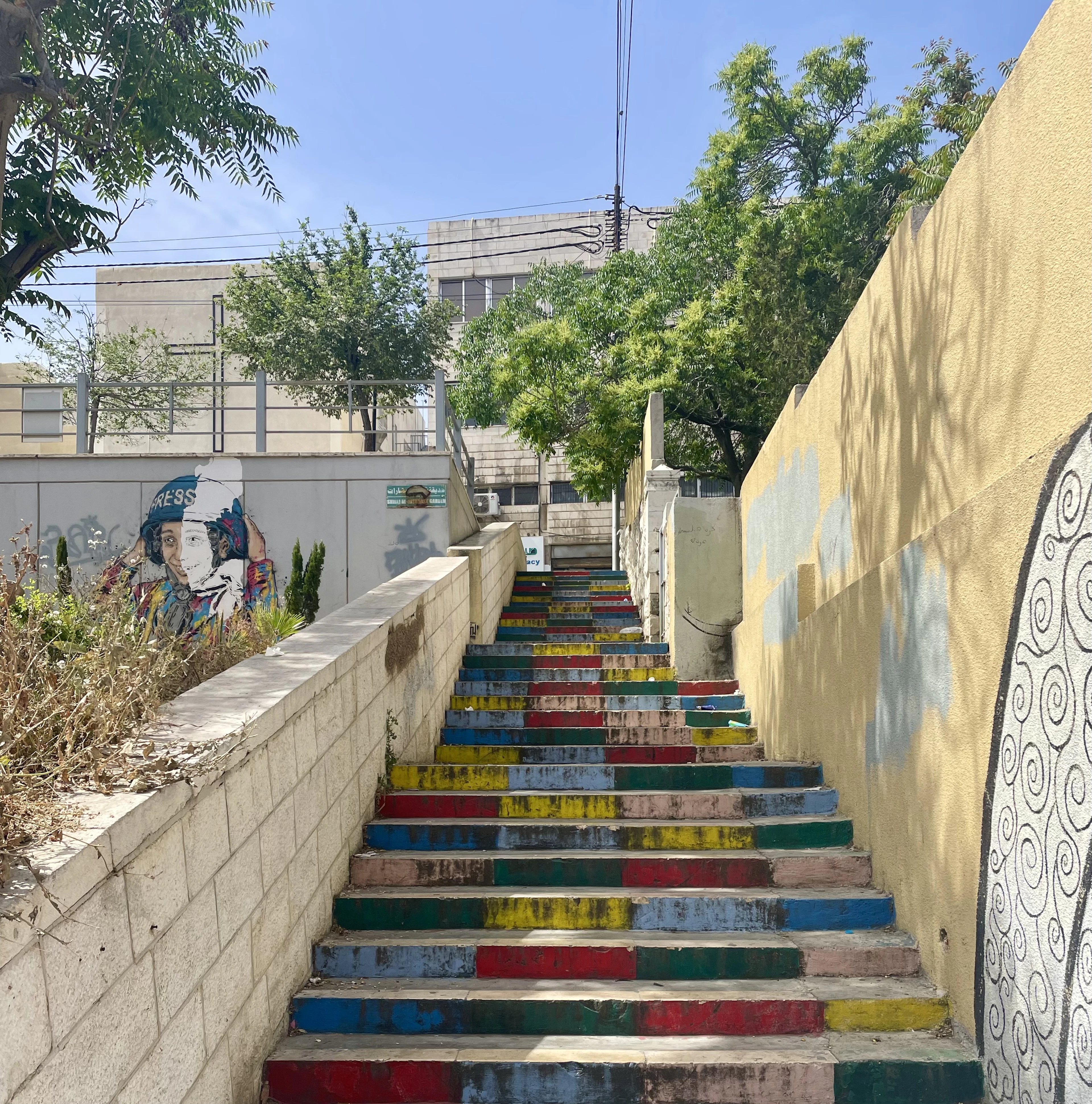 Rainbow-painted stone steps climb past a mural and trees under a blue sky on Amman's Rainbow Street.