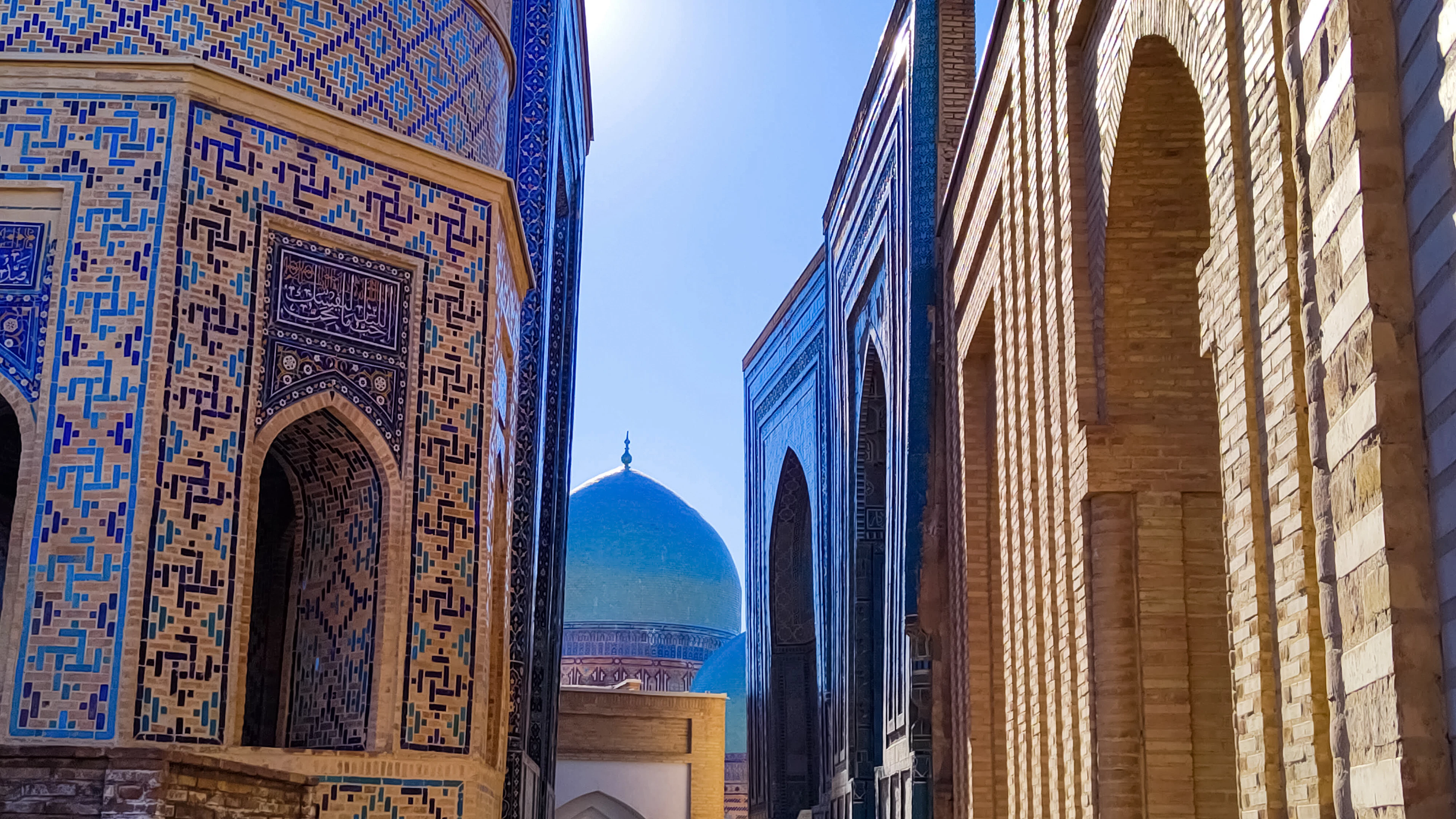 A narrow lane of tiled mausoleum facades at Shah-i-Zinda in Samarkand, a ribbed turquoise dome rising at the end of the passage