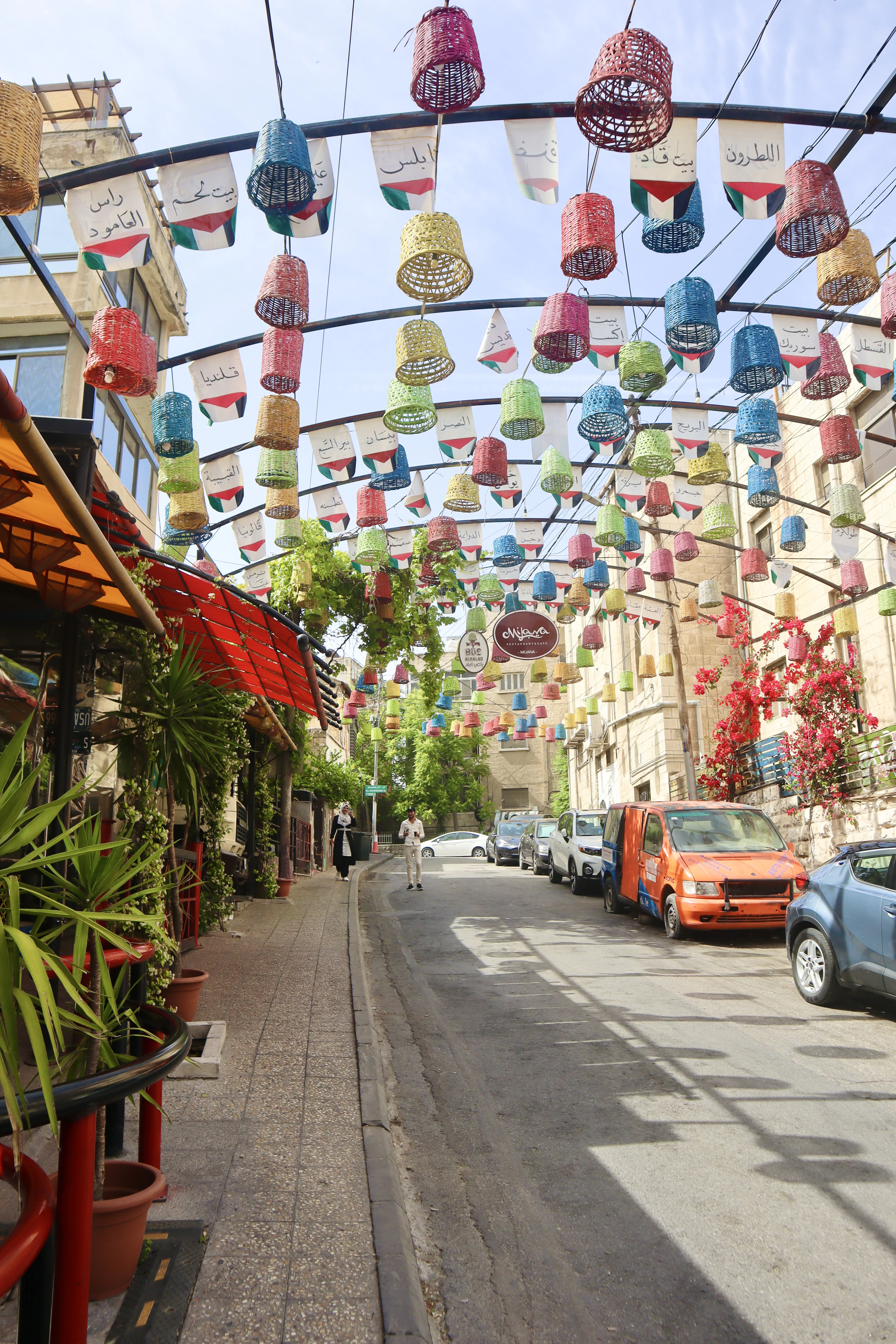 A pedestrian strolls down Amman's Rainbow Street beneath canopies of colorful lanterns and small flags strung between the buildings.