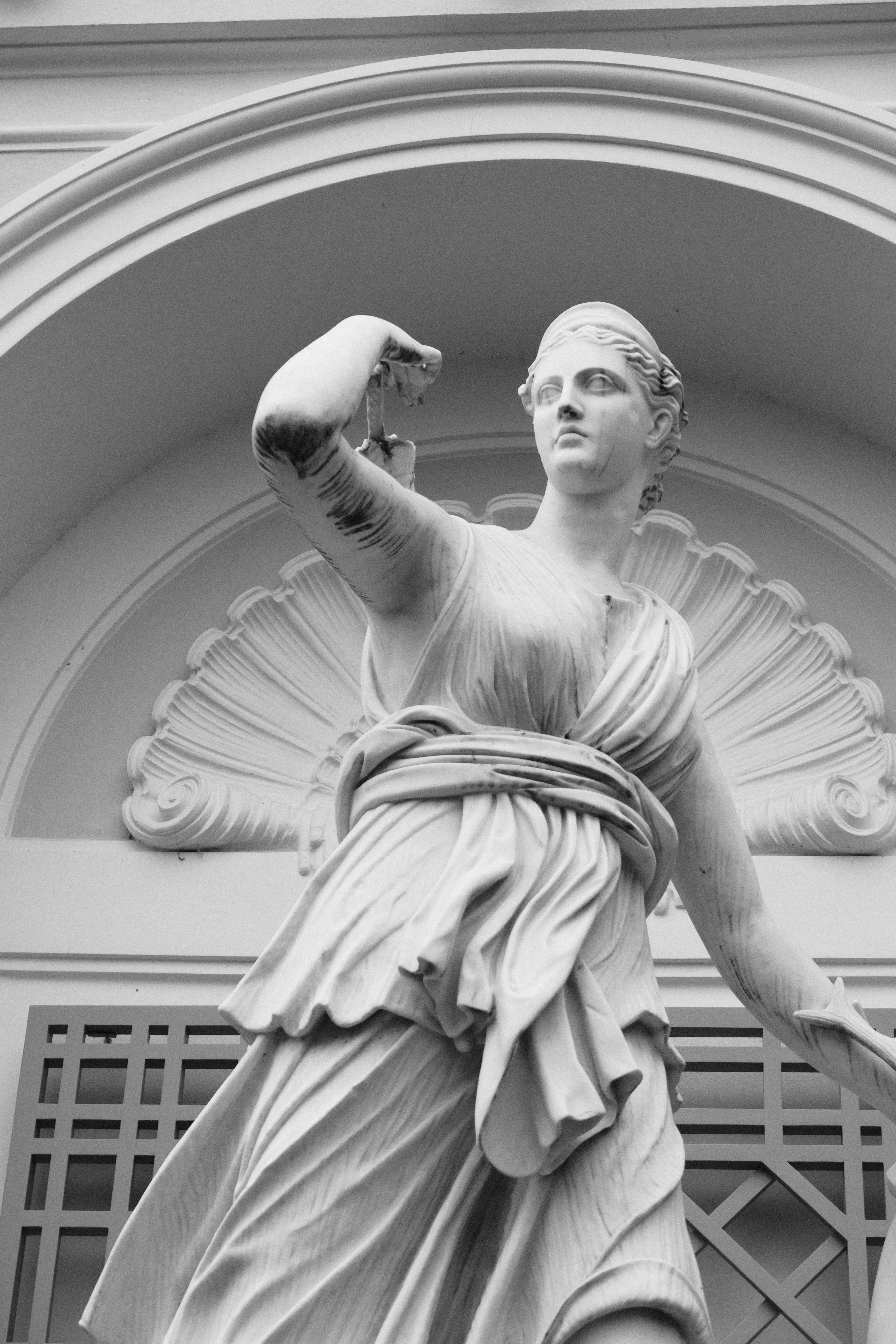 Black and white photograph of a goddess sculpture against architectural backdrop. Dramatic contrast highlighting the sculptural details. Timeless elegance of classical art.
