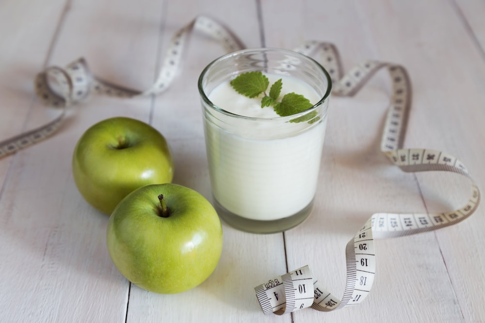 a glass of milk next to two green apples and a measuring tape