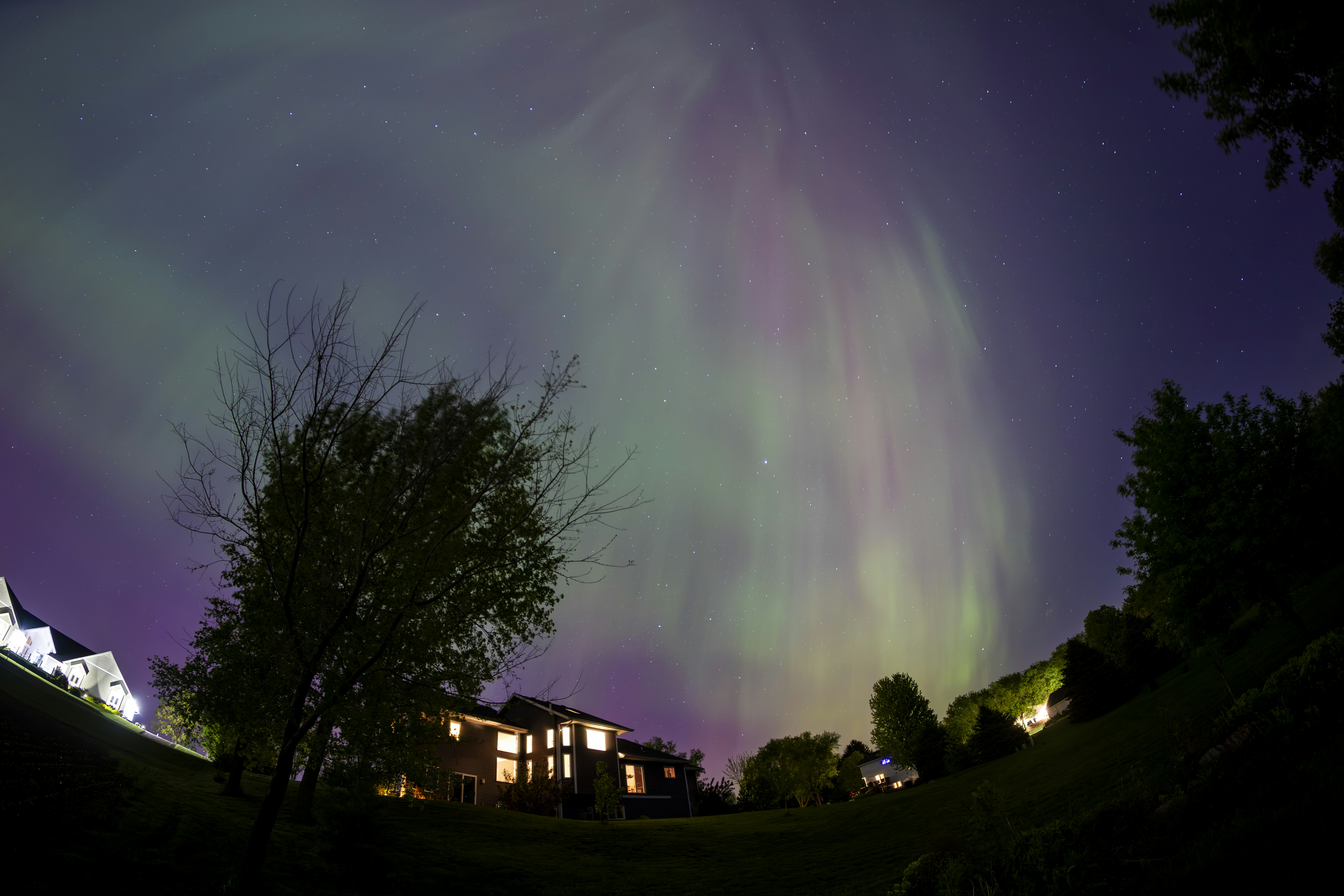 the aurora bore is visible in the night sky, A mesmerizing display of the aurora borealis illuminates the night sky with vibrant green and purple hues, casting an ethereal glow over a peaceful residential neighborhood framed by silhouetted trees.