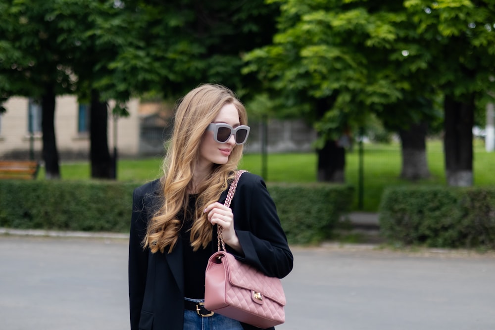 a woman in a black jacket and sunglasses carrying a pink purse