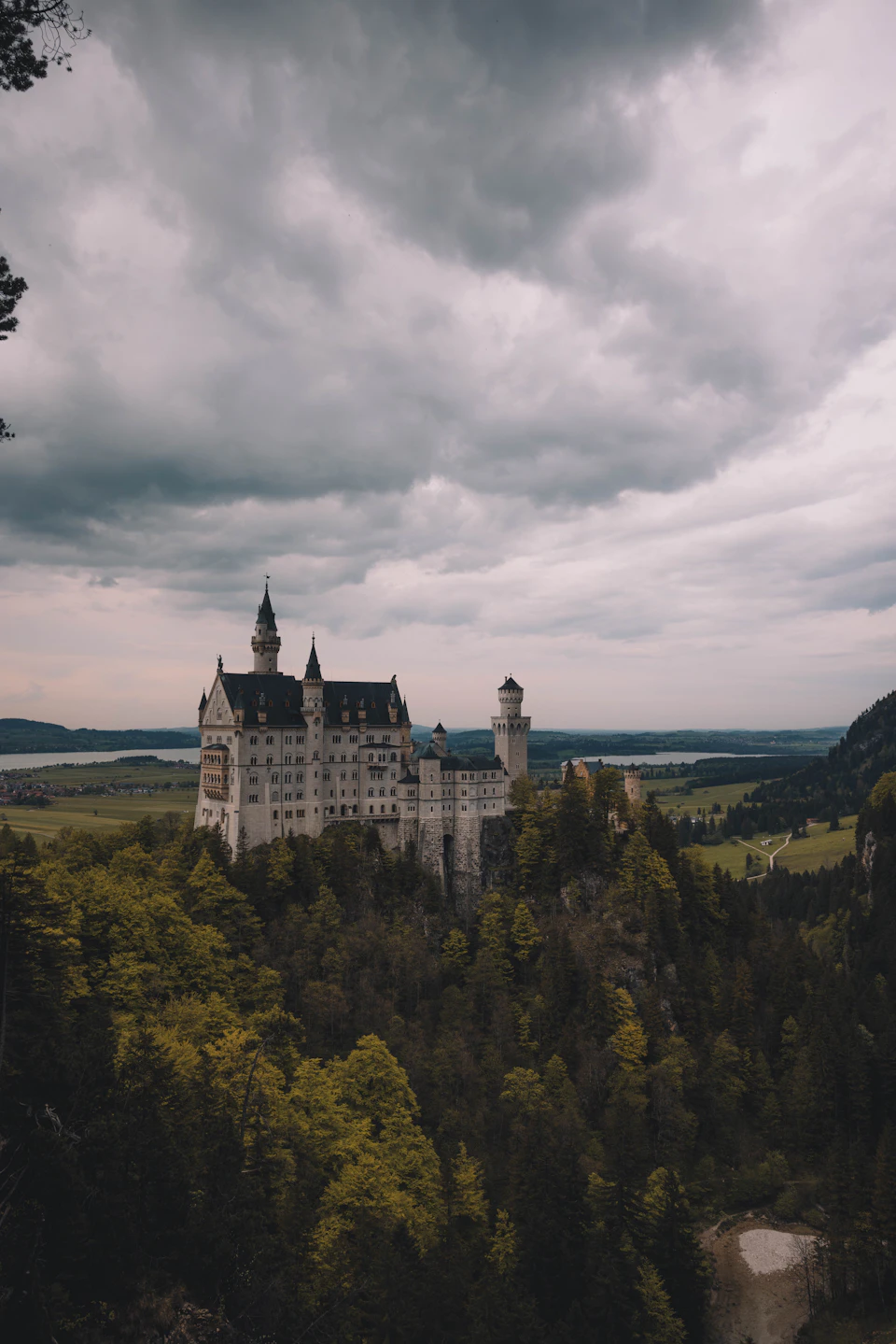 The image depicts a majestic castle perched atop a cliff, overlooking a valley below. The castle is constructed from stone and features multiple towers and turrets that reach towards the cloudy sky. The surrounding landscape includes lush green trees and grassy fields, creating a picturesque backdrop for the historic structure.