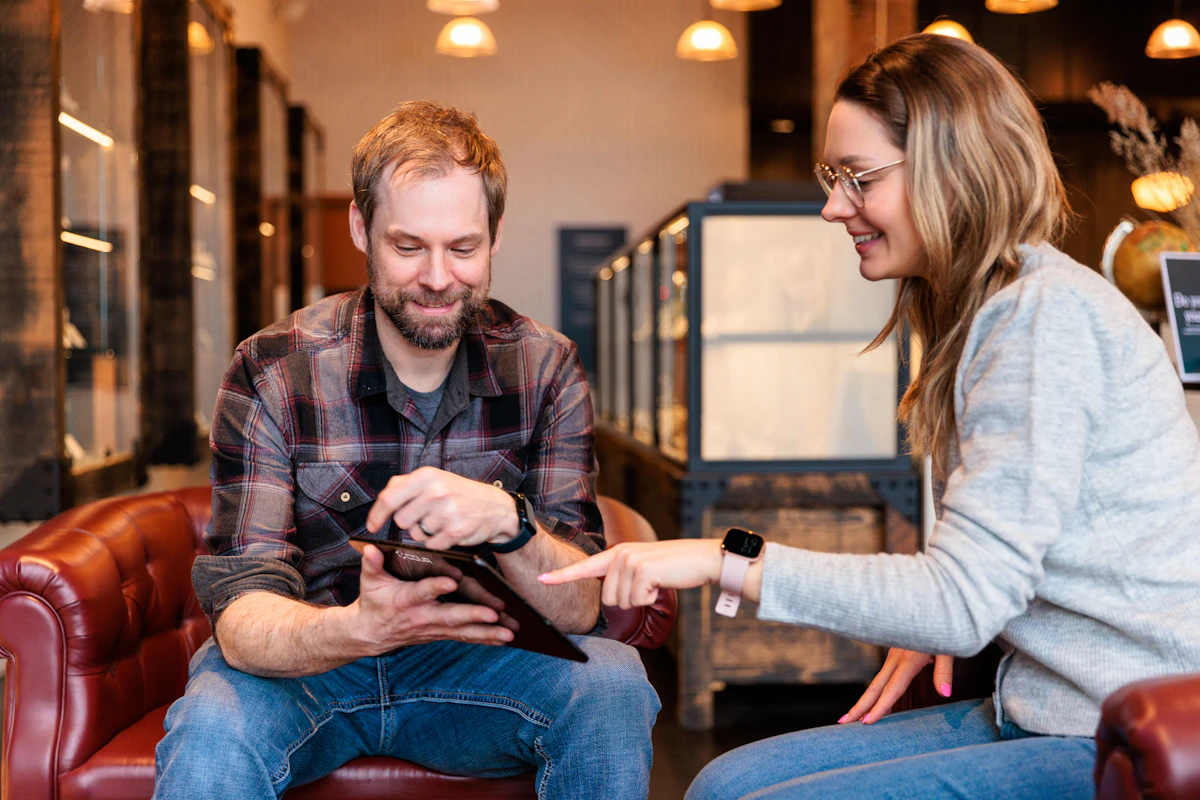 A man and a woman laughing over a tablet on a red leather sofa, her finger on the screen