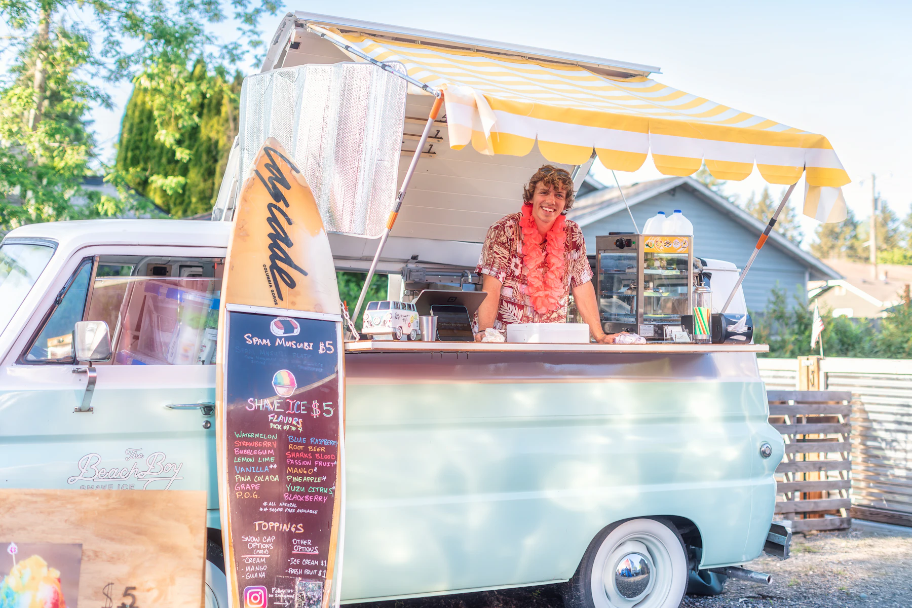 A vintage pale green shave ice truck with a striped awning and a surfboard flavor menu