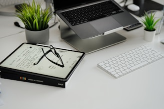 a desk with a laptop, keyboard, glasses and a potted plant