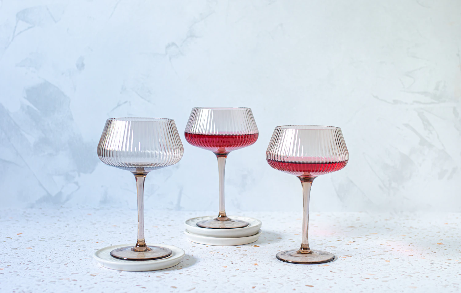 Three ribbed coupes on blush stems, two filled with a pink drink, against a pale wall