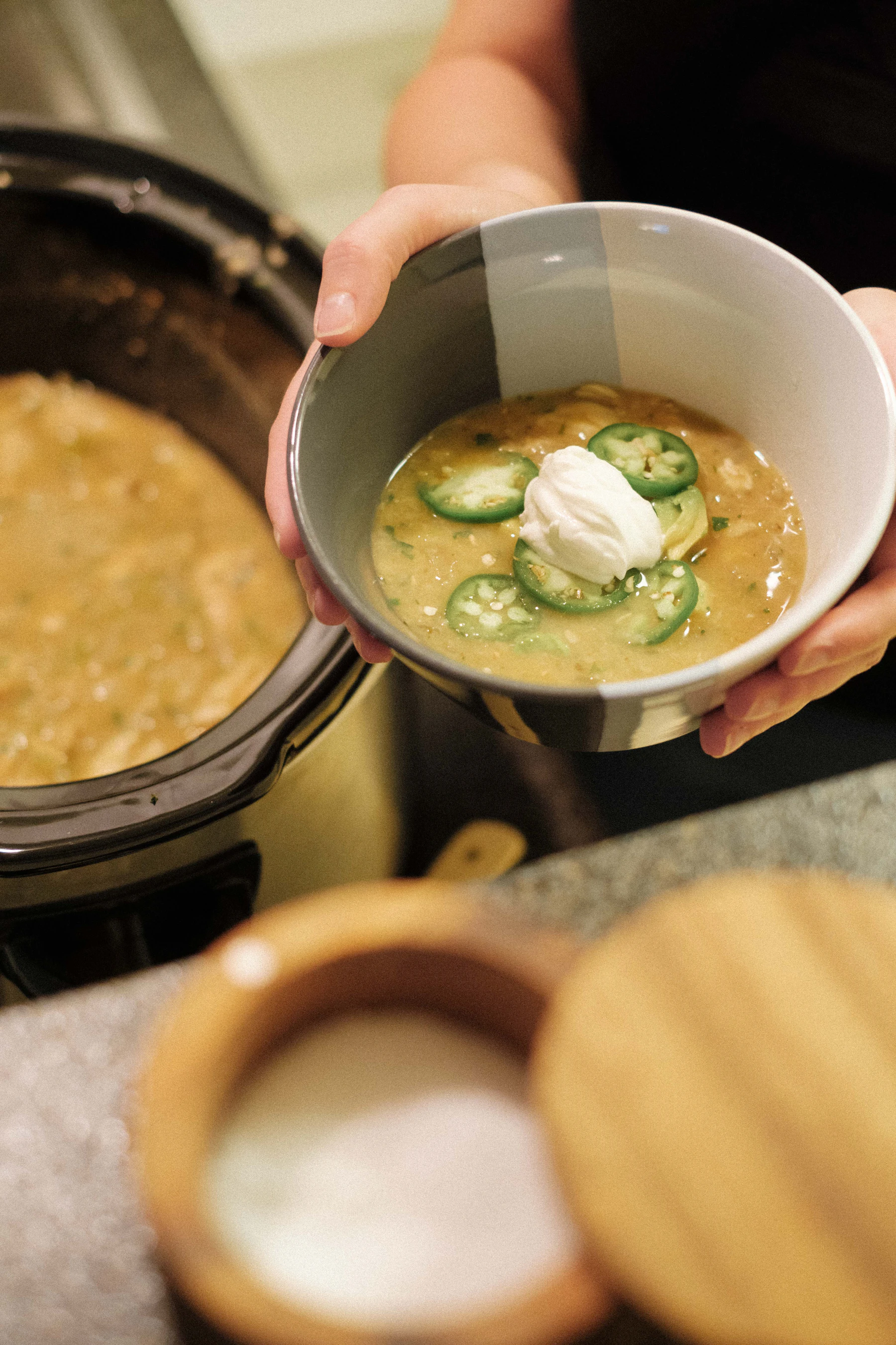 Hands holding a bowl of white chicken chili topped with jalapeño slices and sour cream beside an open black slow cooker