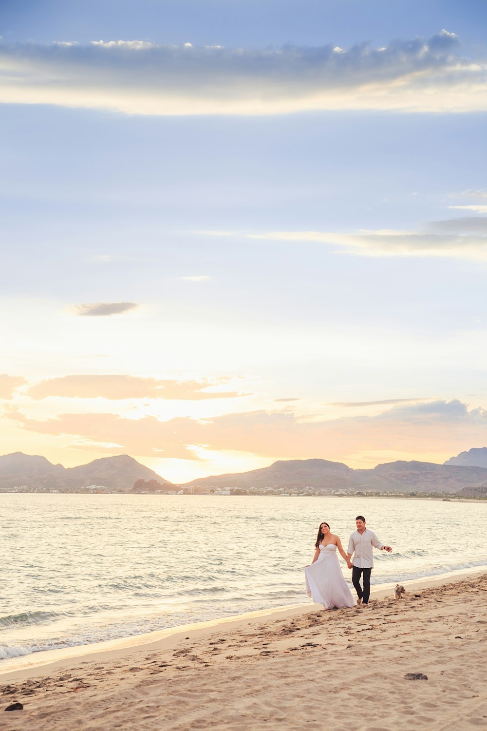 a bride and groom walking on the beach at sunset