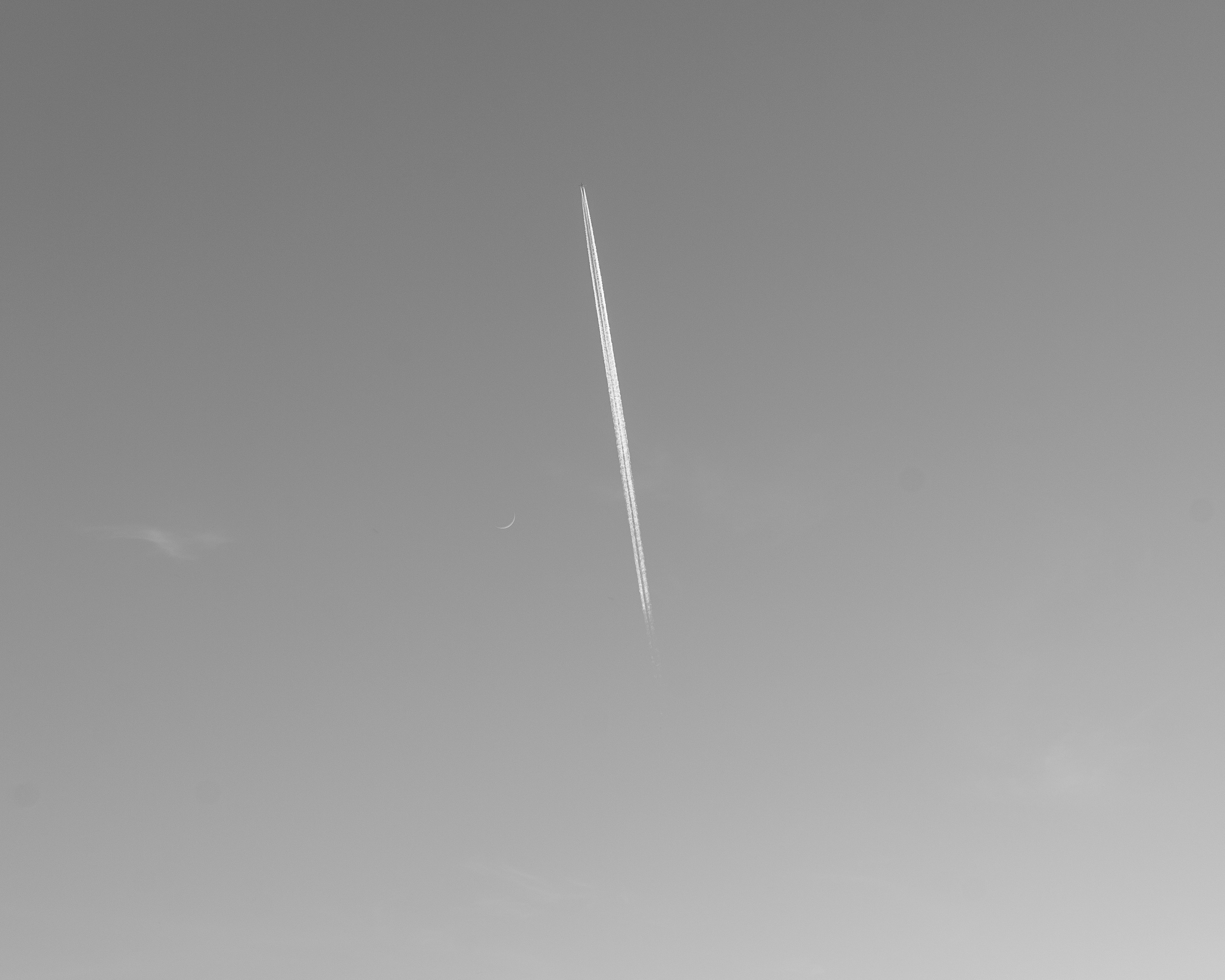 an airplane is flying in the sky with a contrail, A minimalist black and white photograph depicting an airplane contrail slicing through a plain, light gray sky. The long, thin, solitary white line stands out starkly against the monochrome background, drawing the viewer