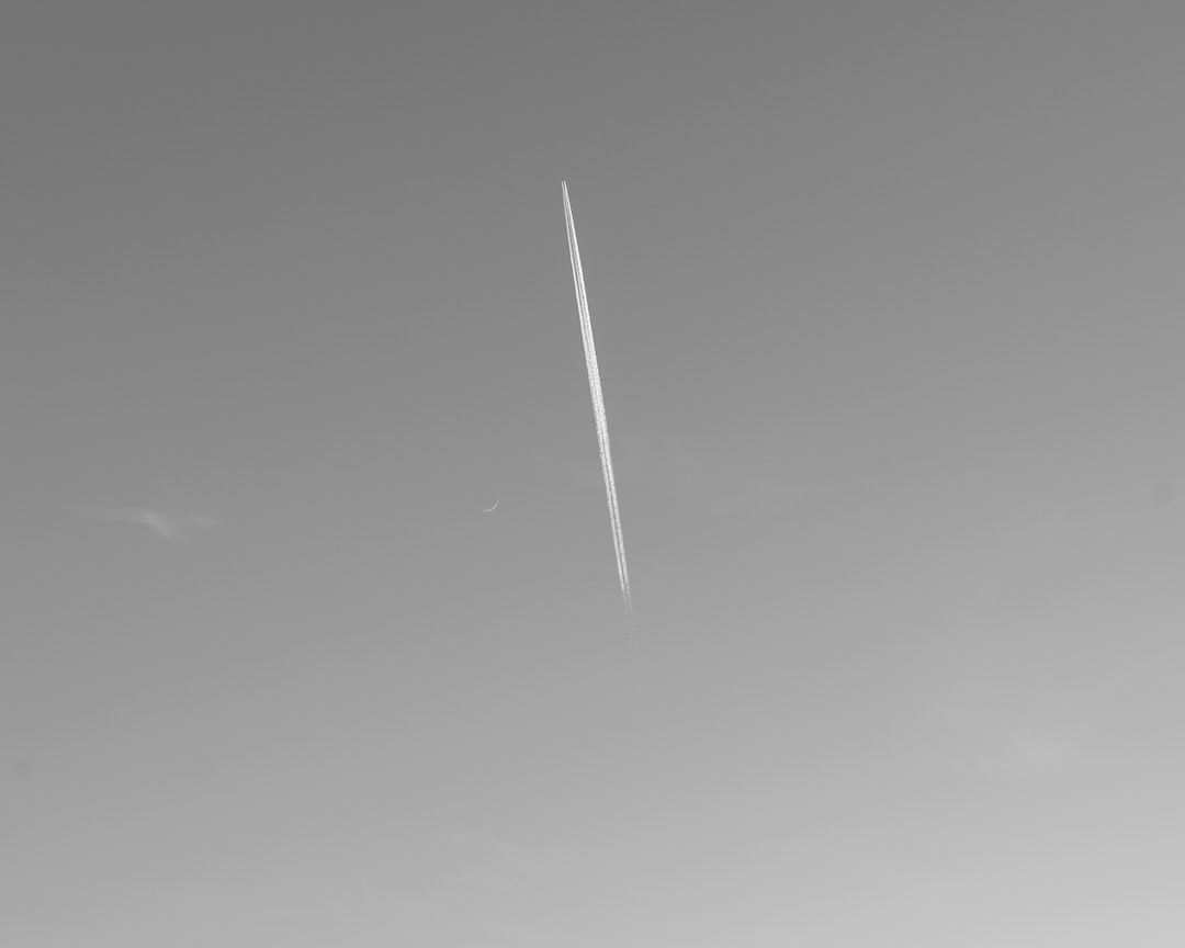 A minimalist black and white photograph depicting an airplane contrail slicing through a plain, light gray sky. The long, thin, solitary white line stands out starkly against the monochrome background, drawing the viewer's eye to the ephemeral trail left behind by the unseen aircraft. This simple striking image evokes themes of travel, solitude, and the fleeting nature of our journeys through the vast expanse of the sky.