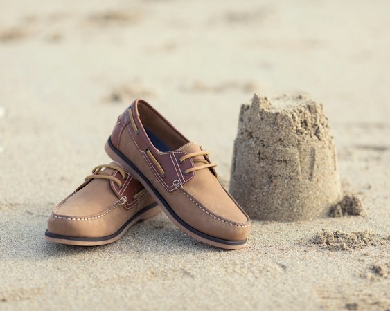 Pair of tan leather boat shoes with rawhide laces and dark soles standing on beach sand beside a small sandcastle