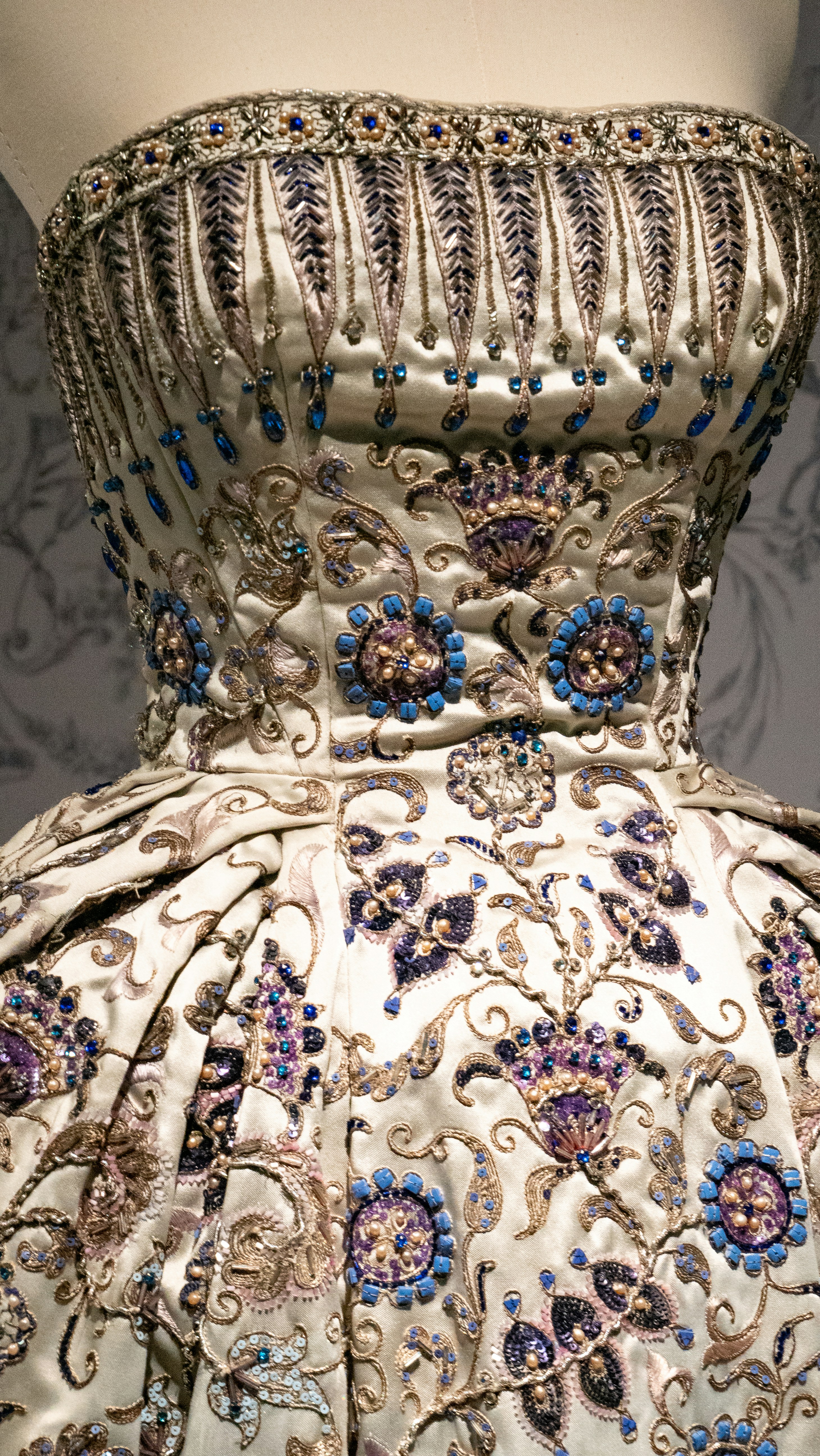 Close-up photograph of an ivory satin dress bodice richly adorned with blue and purple gemstones and gold filigree on a mannequin.