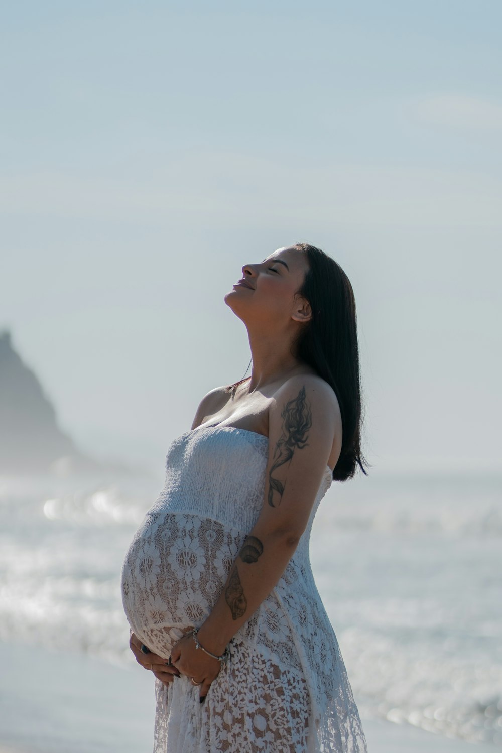 a pregnant woman standing on the beach looking up at the sky