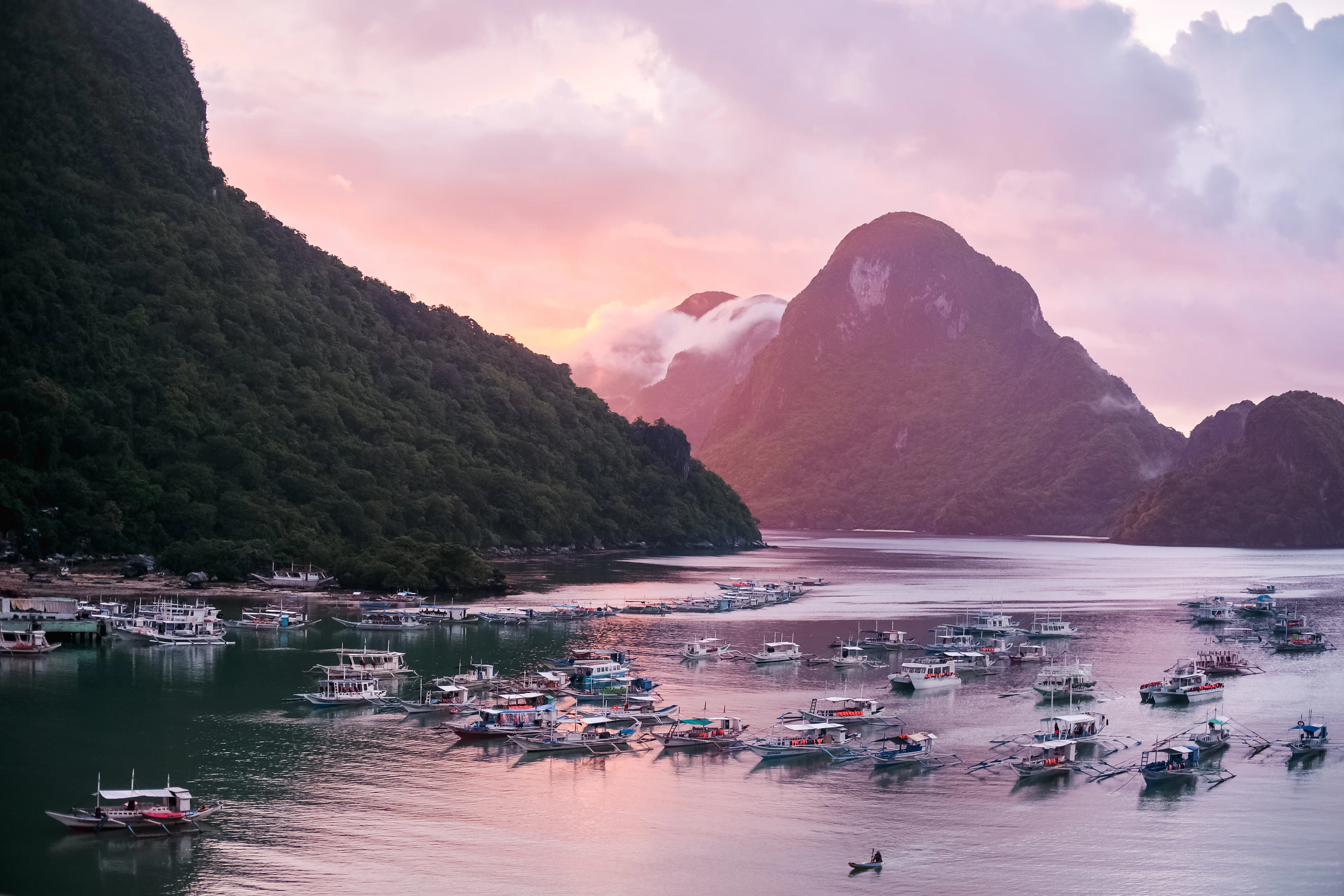 Bangka boats moored in a calm bay beneath limestone cliffs in the pink light of dawn, El Nido