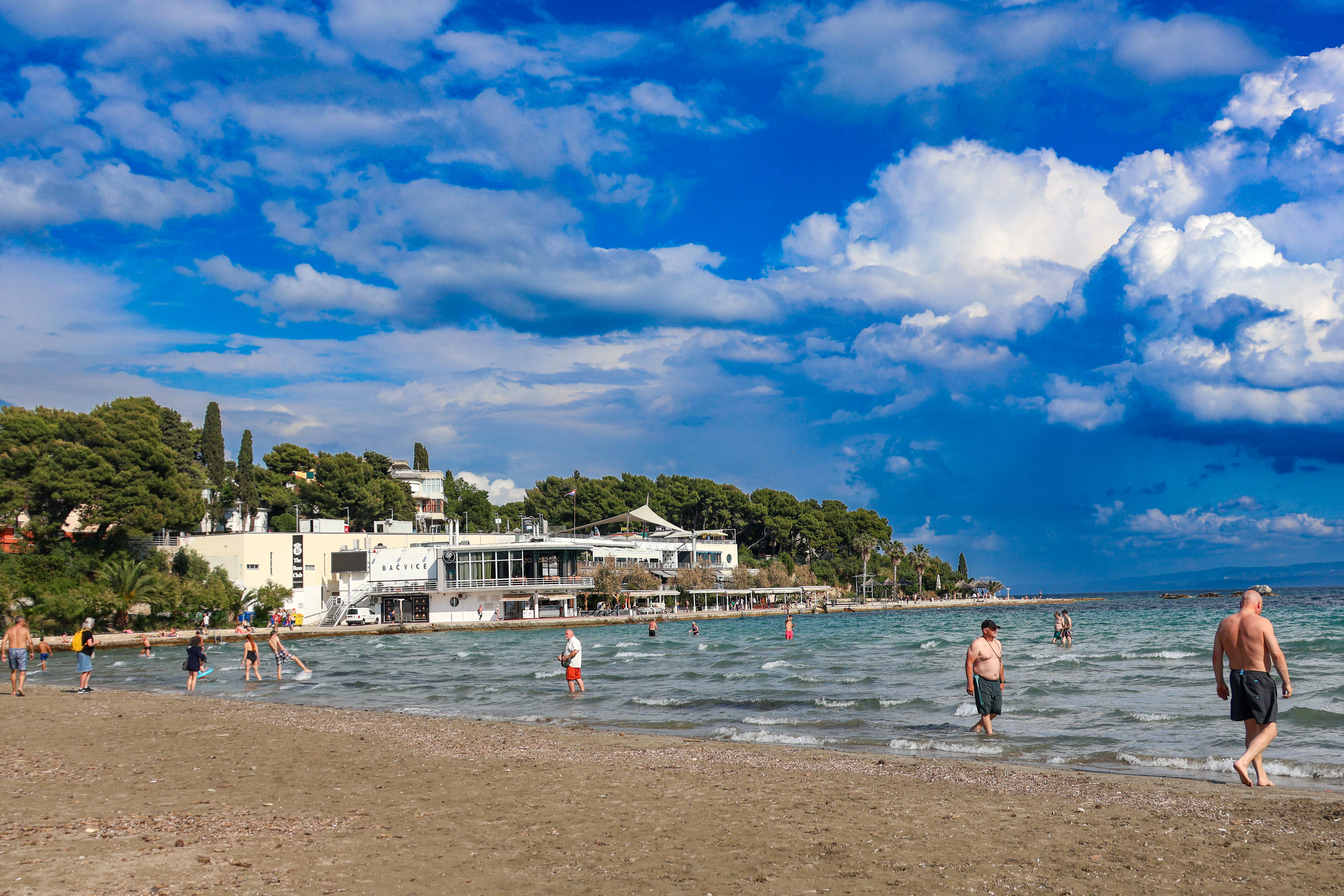 The shallow sandy bay of Bačvice in Split, bathers standing far out in knee-deep water beneath towering white cumulus, with the beach pavilion and its pine-covered slope running along the shore.