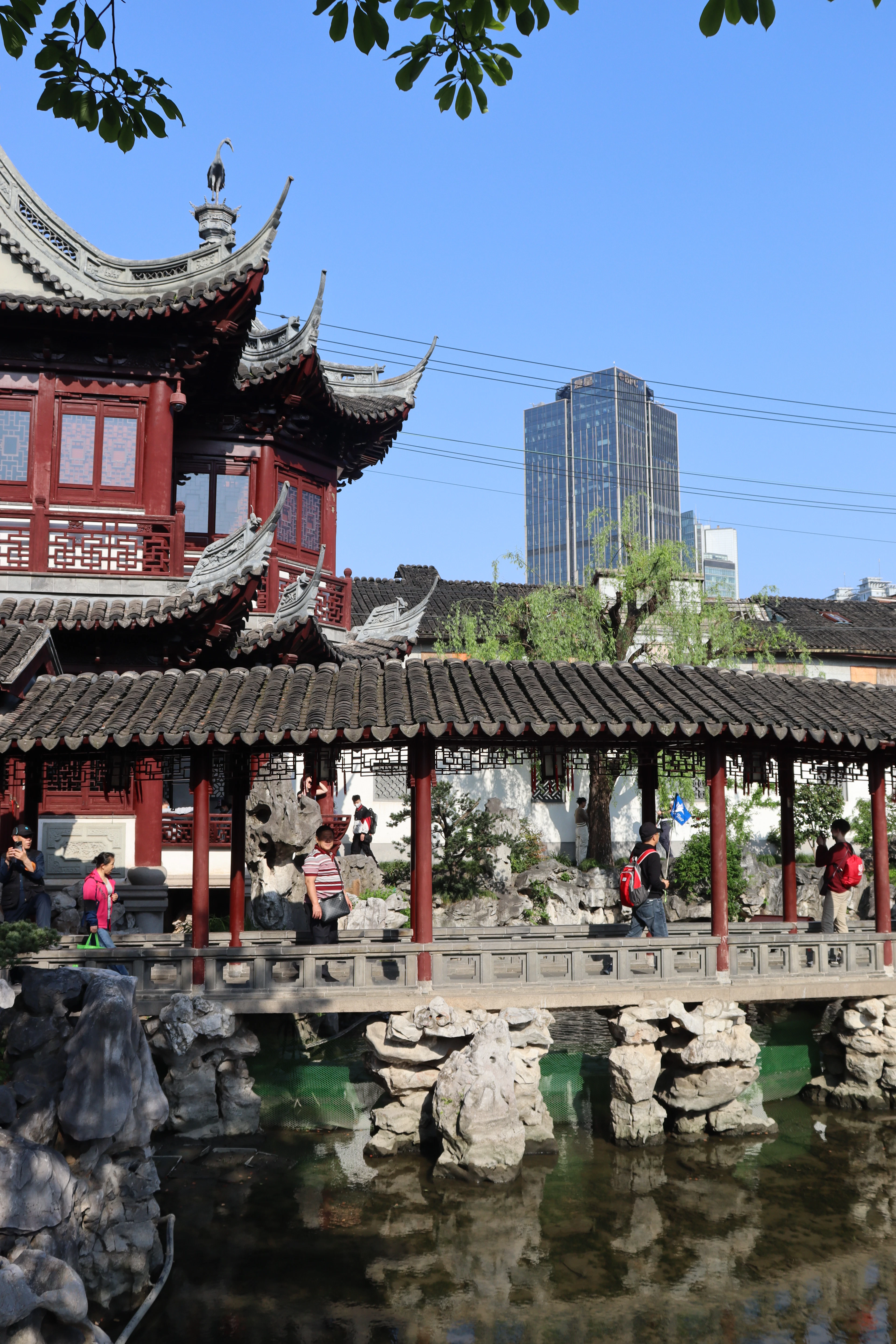 A covered walkway on red columns crossing the rockery pond at Yu Garden in Shanghai, its flying tiled eaves and lattice screens framed against a glass office tower rising just beyond the garden wall.