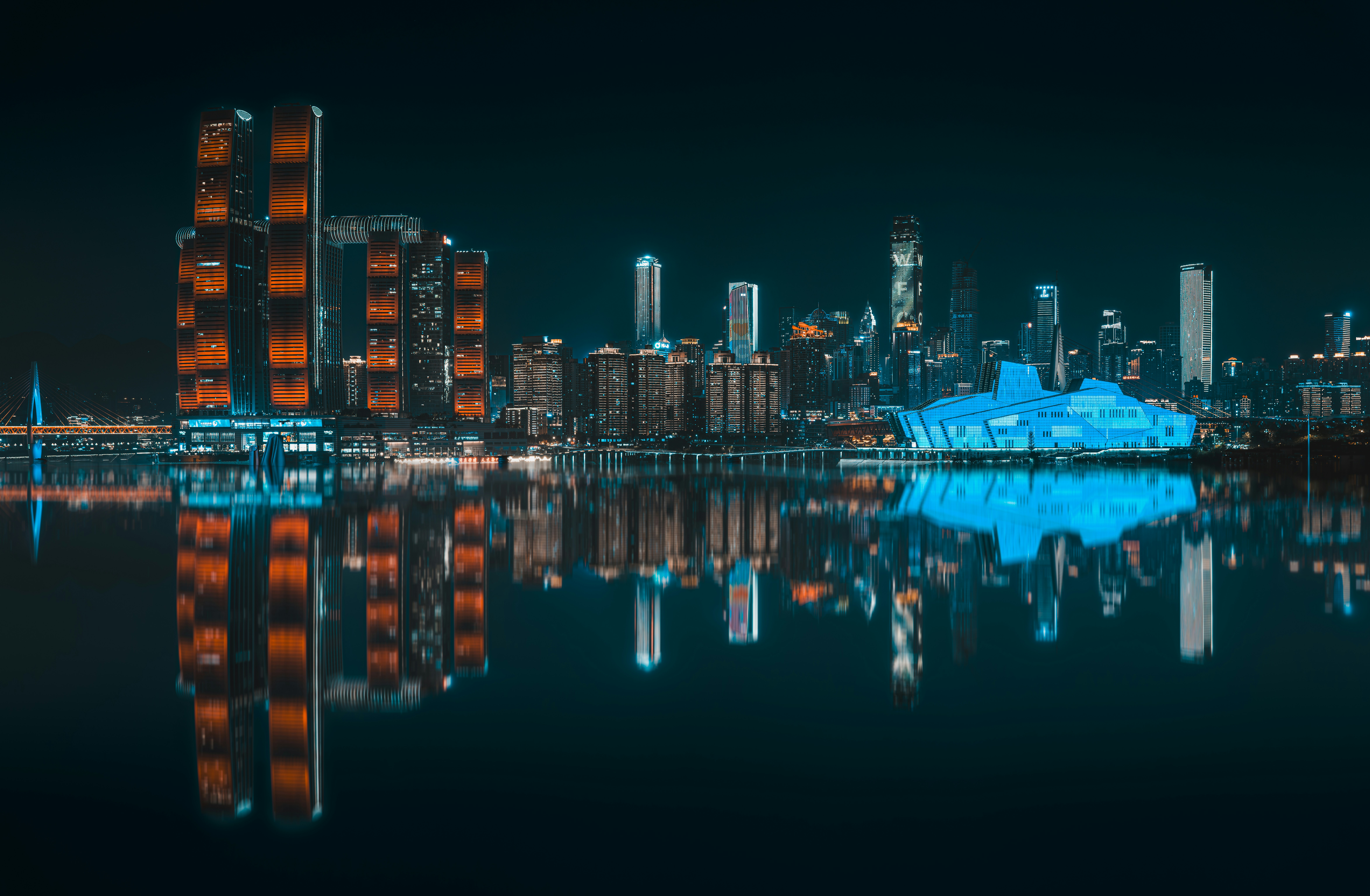 City skyline at night, deep blue darkness, glowing amber and white building lights reflecting in dark water below