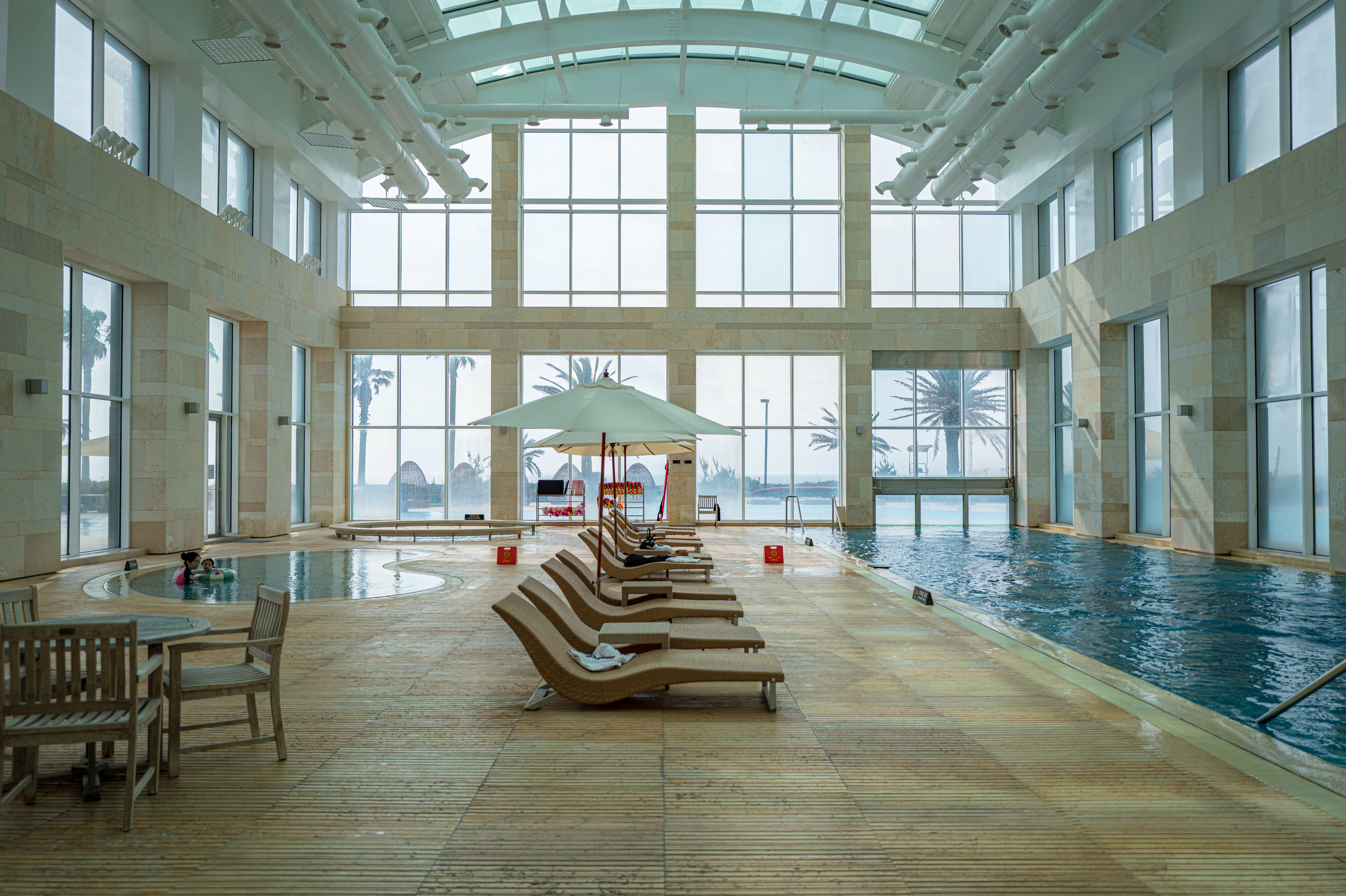 A tall glass-walled indoor pool hall at a resort in Seogwipo on Jeju Island, wooden loungers and a white parasol set out along the water with palm trees visible through the windows.