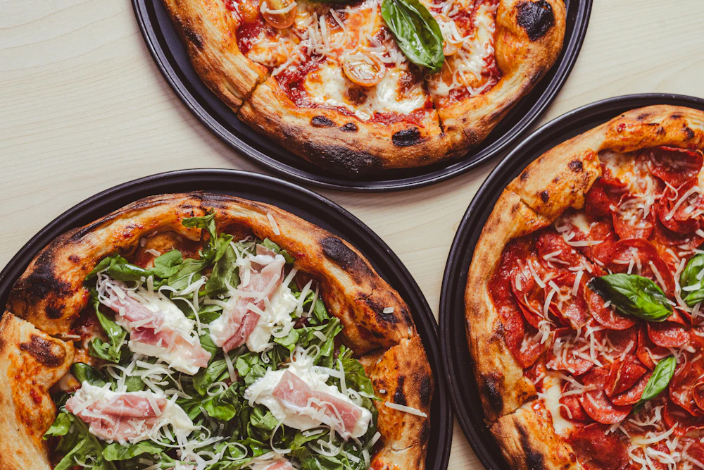 Three pizzas on metal trays seen from above: one with arugula and prosciutto, one with cherry tomatoes and grated cheese, one margherita