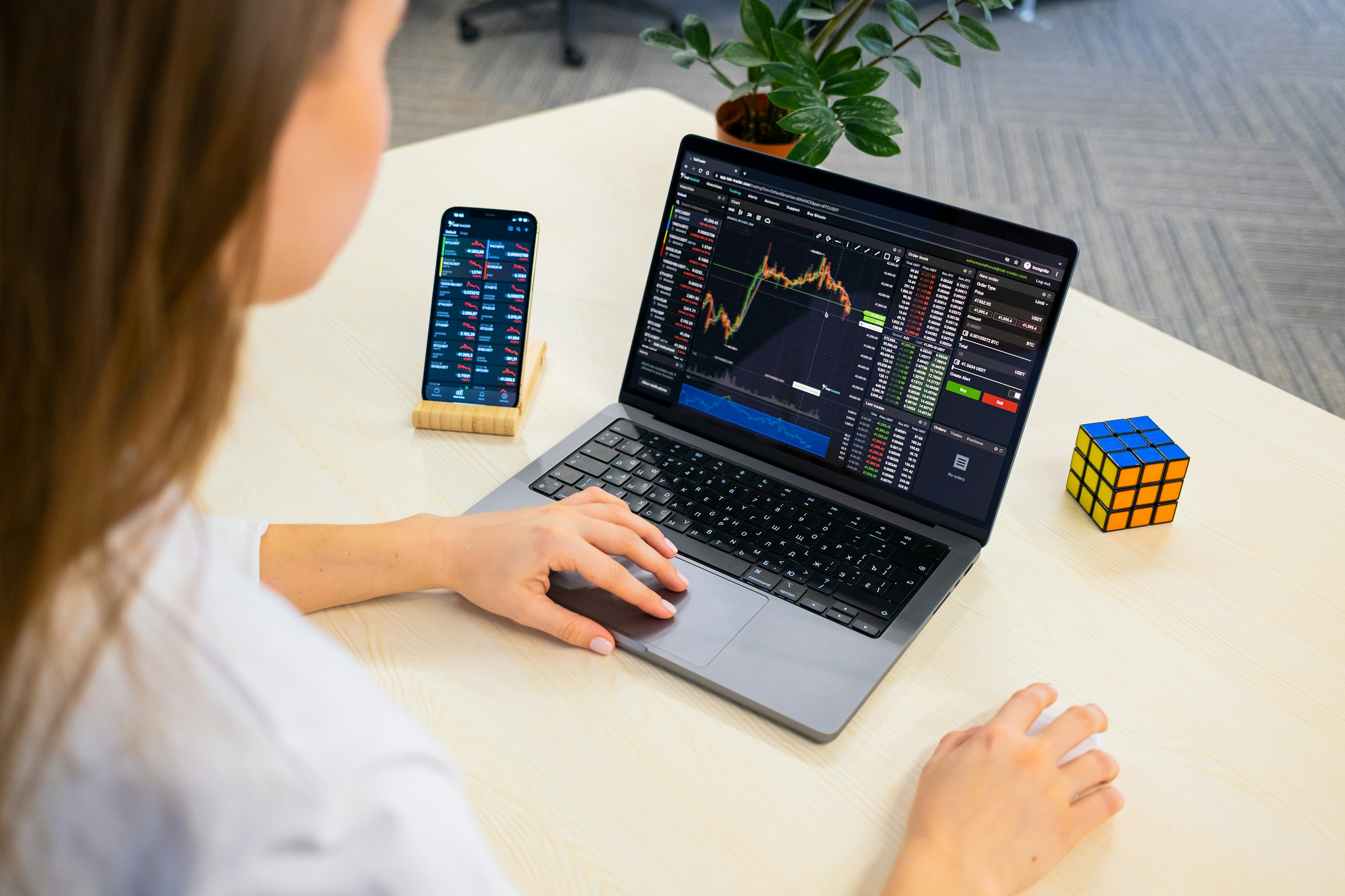 Woman crypto trader investor broker analyst businessman using mobile phone app analytics for cryptocurrency financial market analysis, chart graph index on smartphone and computer. Over shoulder view.