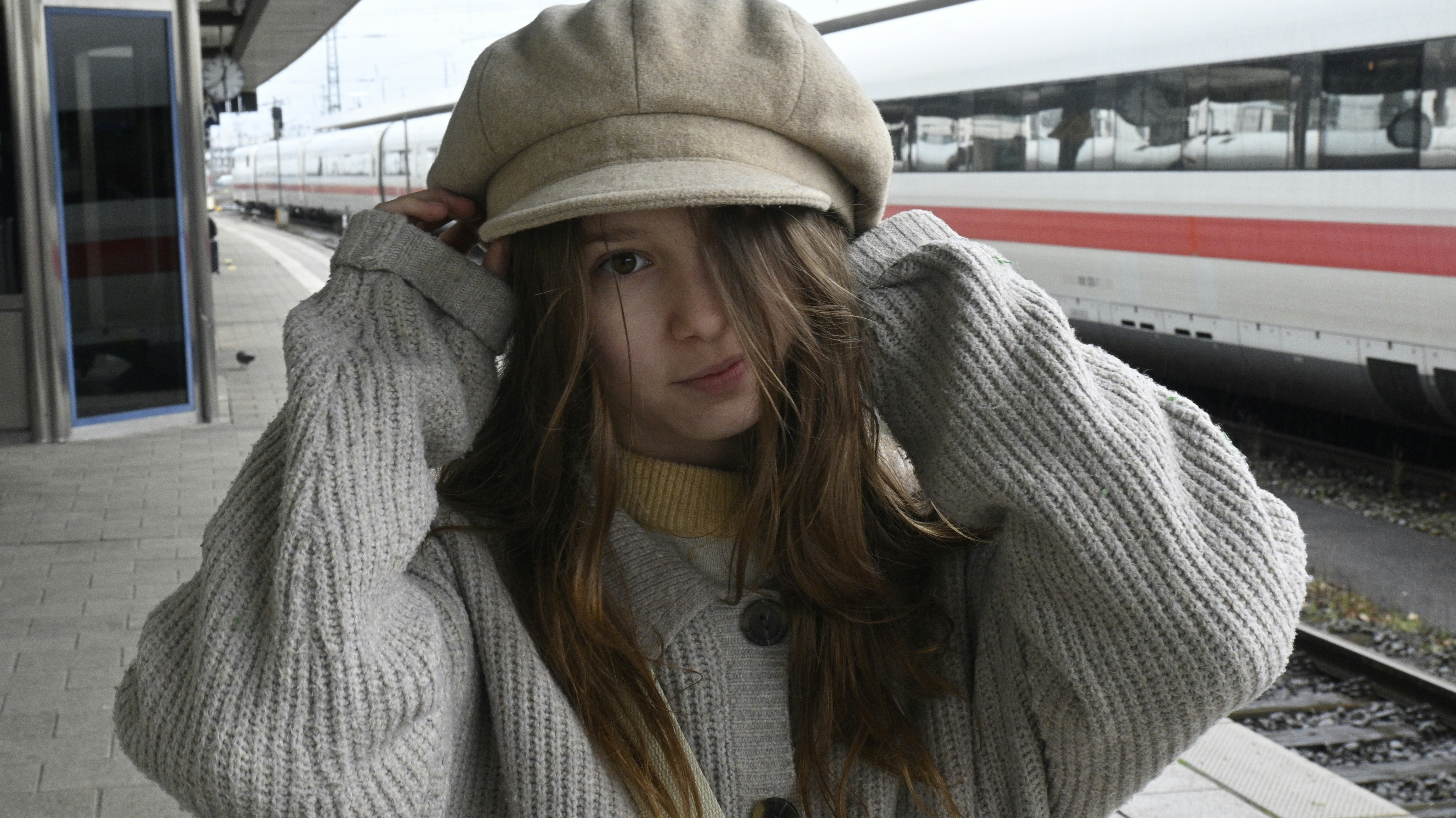 a girl in a gray sweater and a hat stands in front of a train