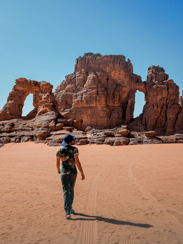 a man walking across a sandy field next to a rock formation