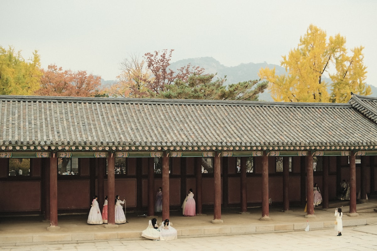 A group of people wearing traditional Korean hanbok in pastel colors of white, light pink, and lavender stand before a long palace building with dark tiled roof and wooden pillars.. Photo by Austin Curtis on Unsplash.