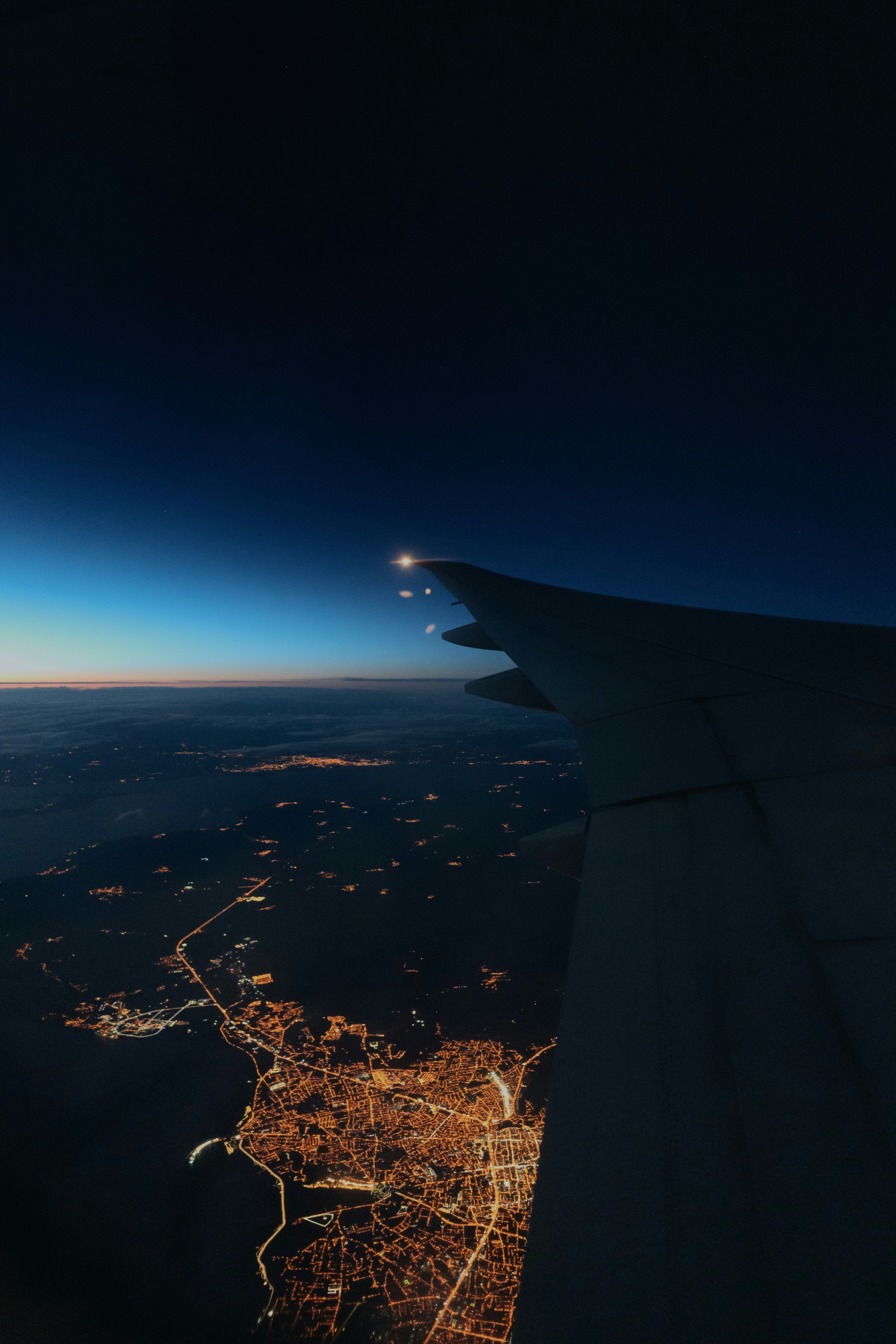 City lights twinkle beneath a wing as twilight descends, creating a serene transition between day and night.